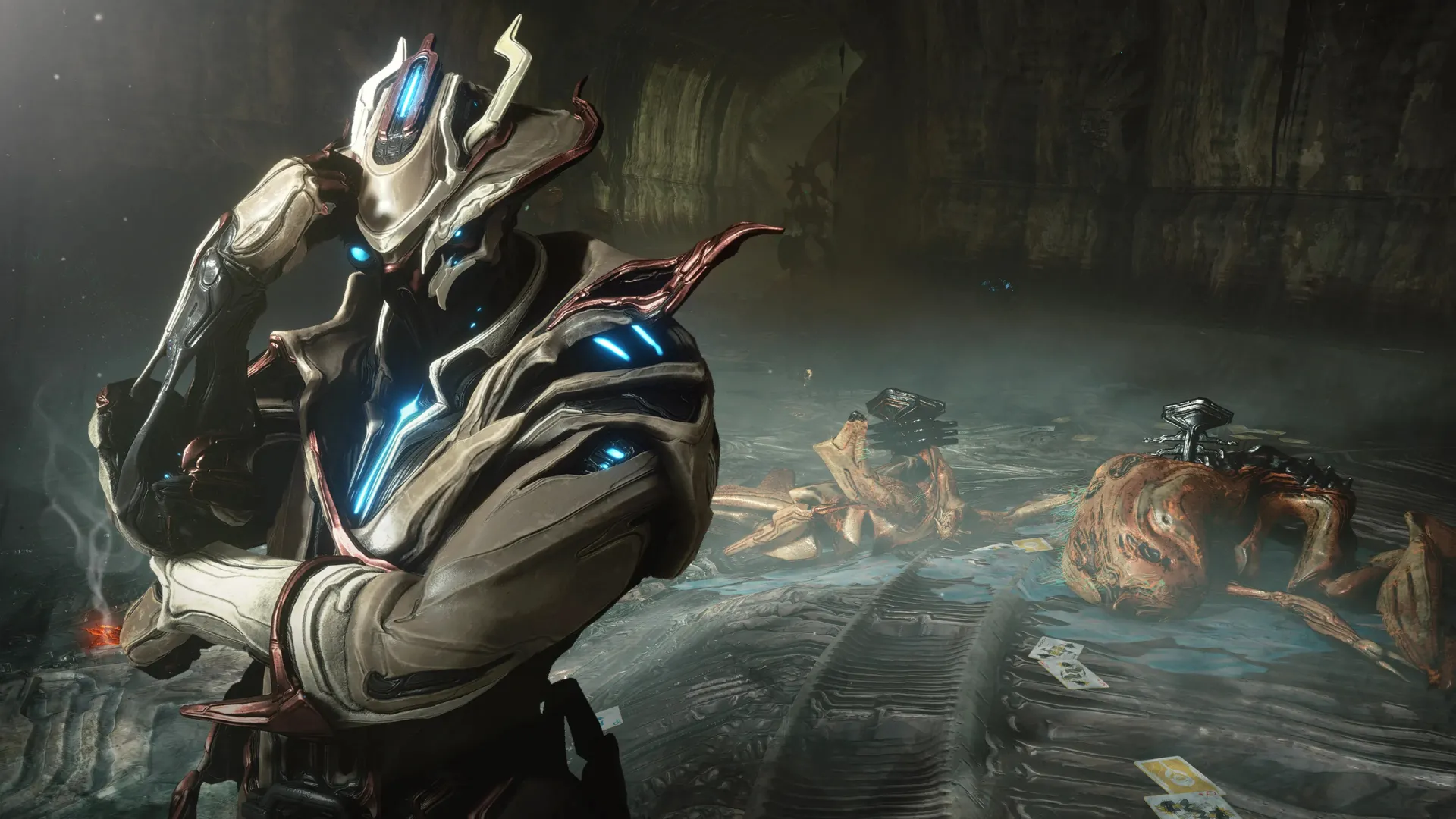 A Warframe stands in a dim cave beside defeated enemies, with playing cards scattered across the wet ground.