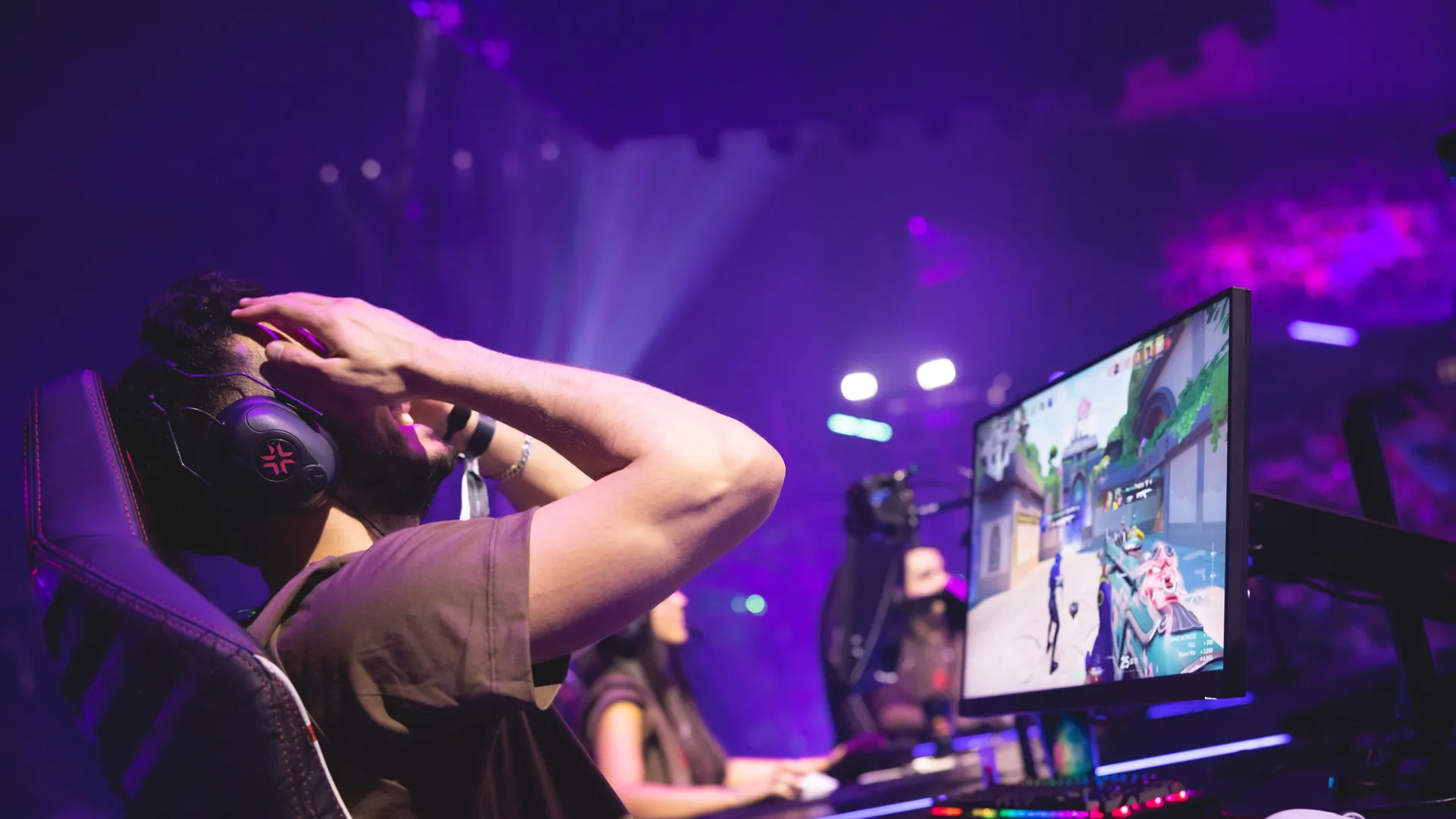A player in a headset throws his hands to his head reacting to Valorant on a monitor at a purple-lit esports stage.