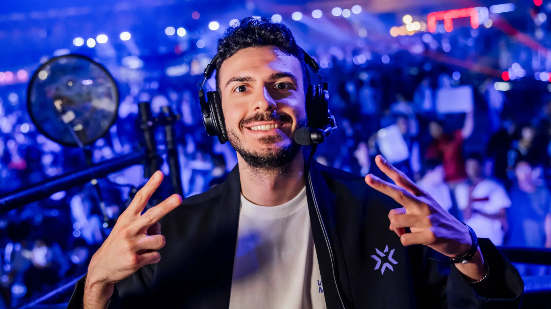 Tarik grins and makes peace signs with both hands, wearing a headset and dark jacket at a blue-lit Valorant event.