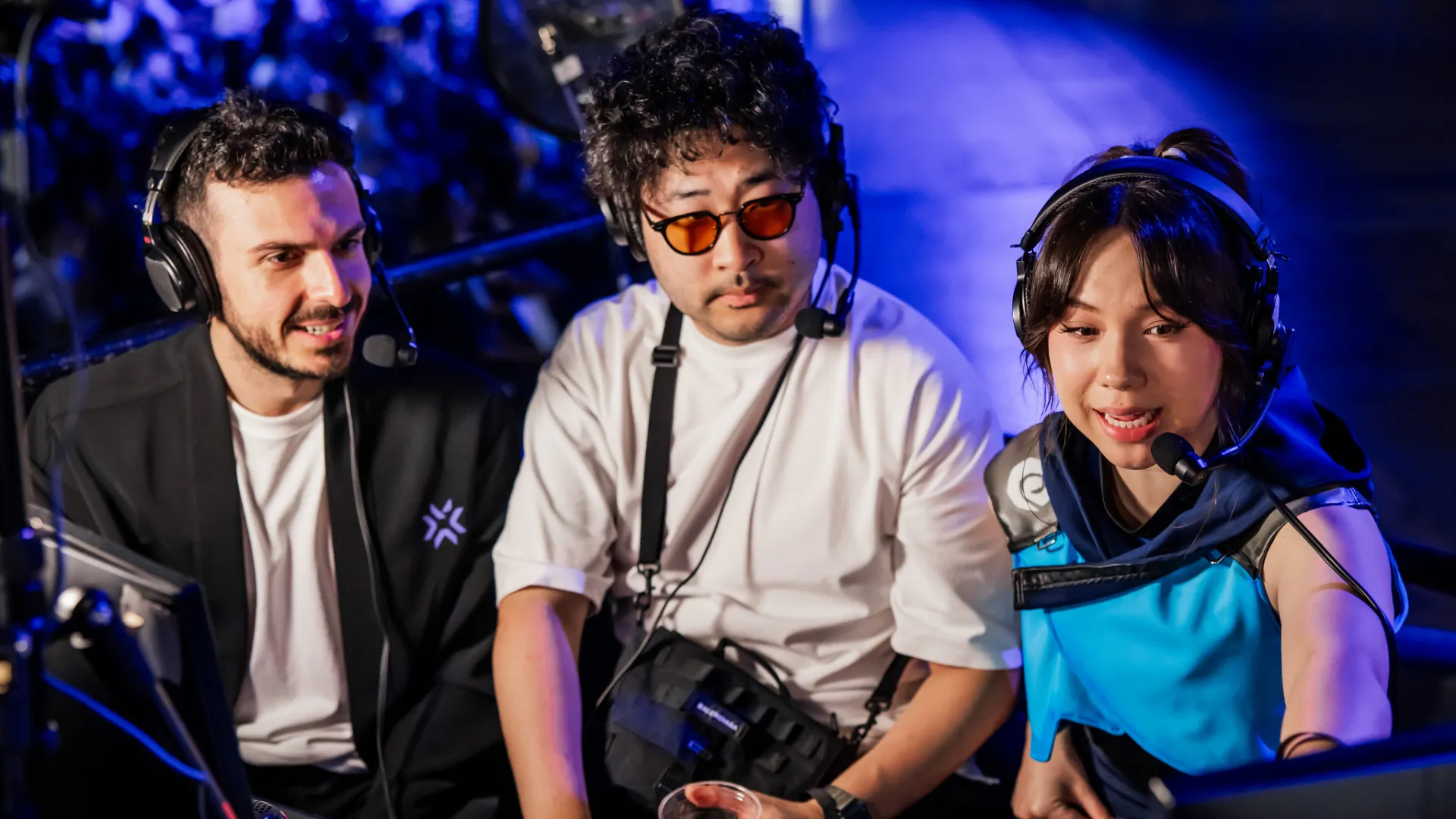 Tarik smiles beside two other people in headsets at a blue-lit Valorant event, one wearing a bright blue jersey.