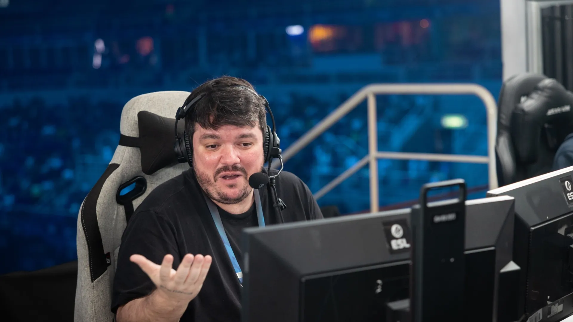 Gaules gestures with one hand while casting into a headset mic at an ESL Counter-Strike event, arena seating behind him.