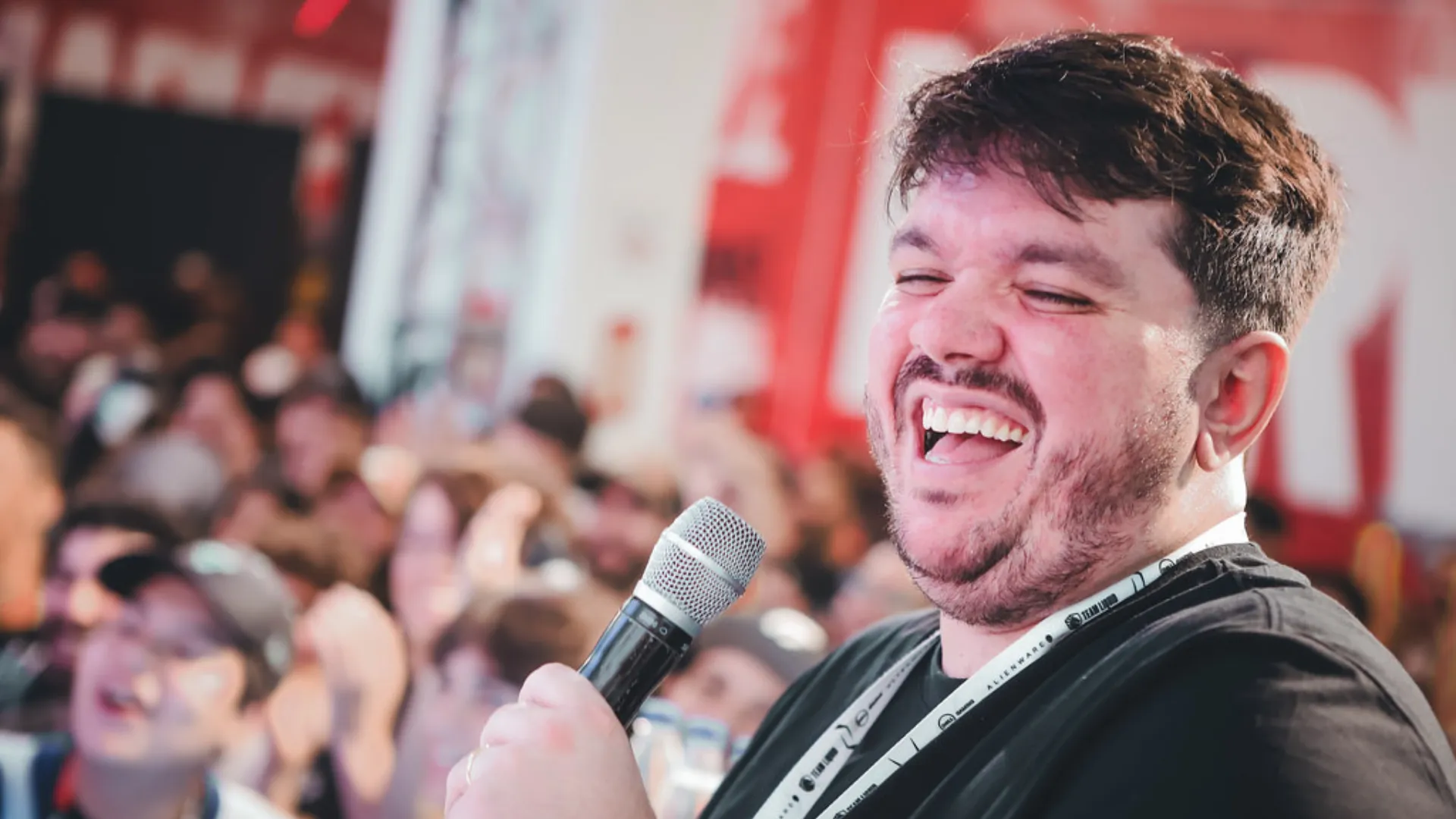 Gaules laughs with his eyes shut, holding a microphone in front of a blurred crowd at a live esports event.
