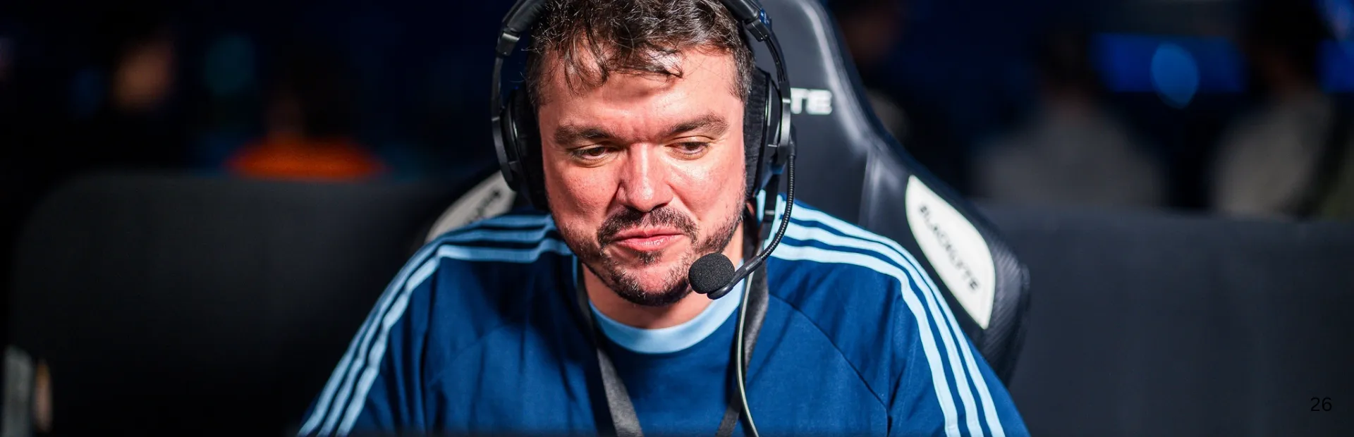 Gaules looks down in concentration wearing a blue jersey and headset while casting at an esports event.