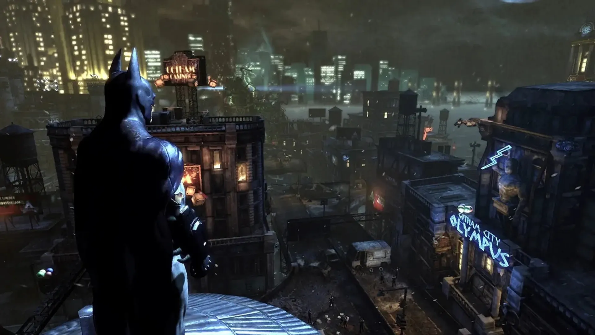 Batman stands on a rooftop overlooking the neon-lit streets and towers of Gotham at night in Batman: Arkham City.