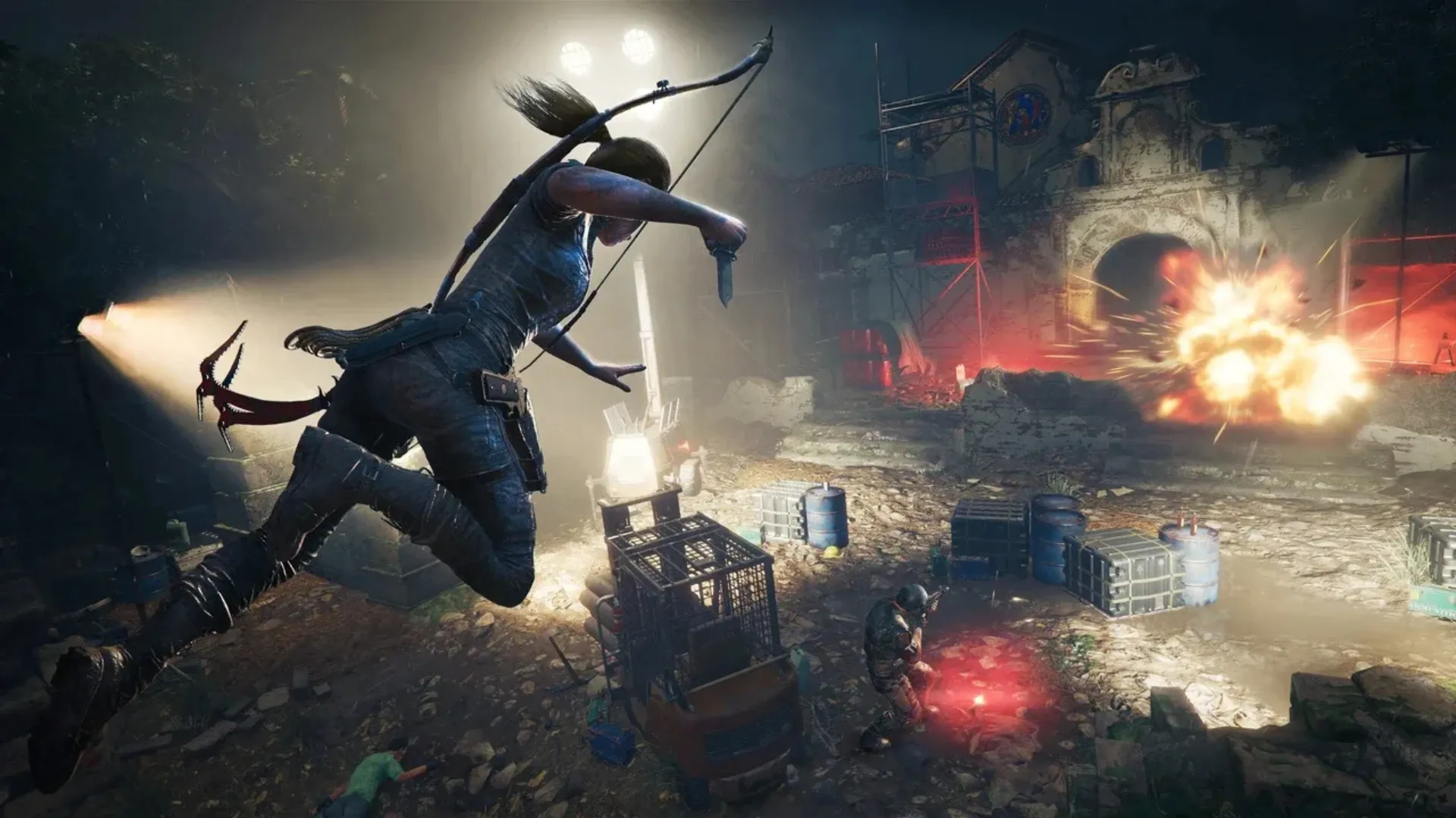 Lara Croft leaps mid-air with a bow on her back as an explosion erupts across a floodlit enemy camp at night.
