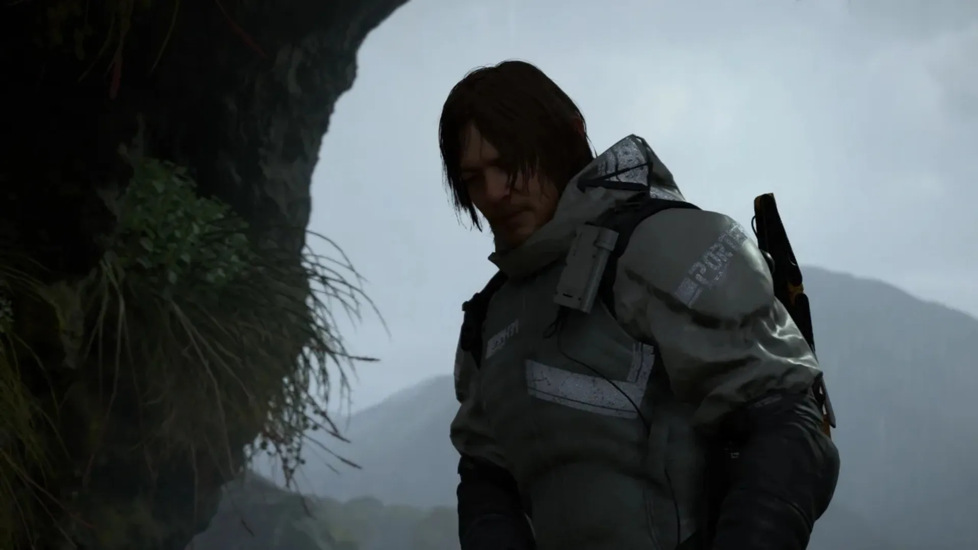 Sam Porter Bridges stands with a loaded backpack beside a mossy rock face under a grey, rainy sky in Death Stranding.