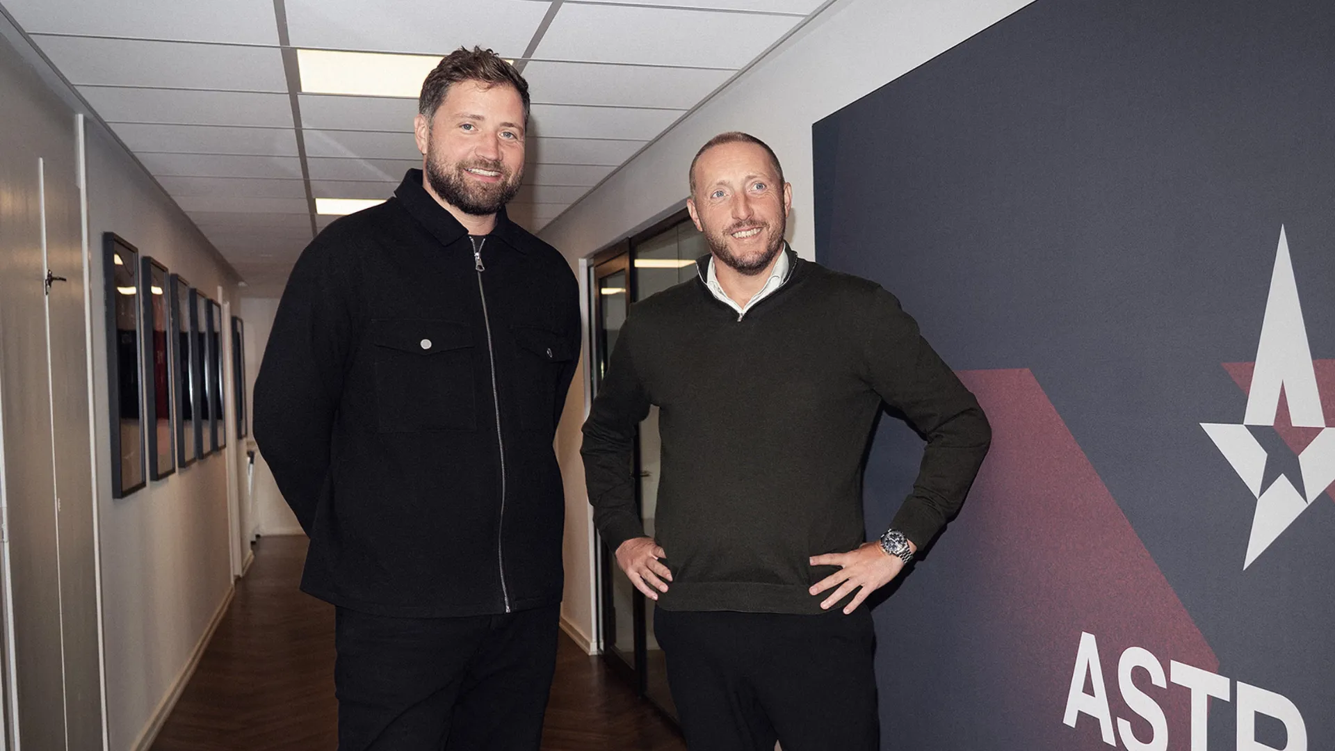 Two men in dark clothing smile in an office hallway beside a wall bearing the red and white Astralis star logo.