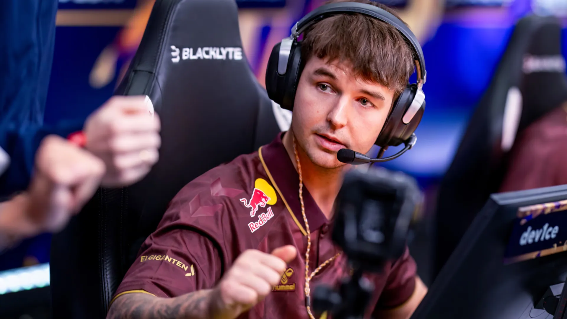 A player in a maroon Astralis jersey and headset speaks at a monitor labeled device during a live CS2 match.