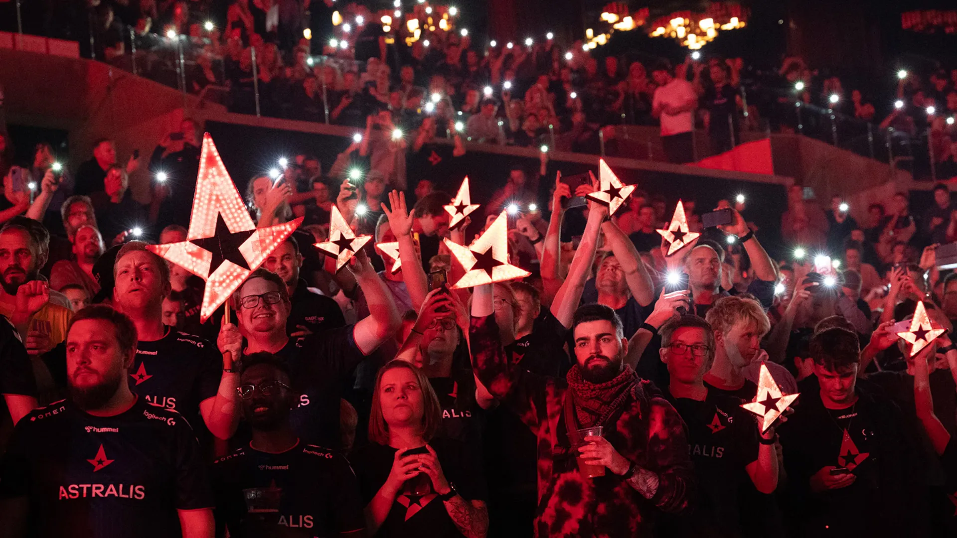 A red-lit crowd holds up glowing Astralis star signs and phone lights at a packed Counter-Strike arena.