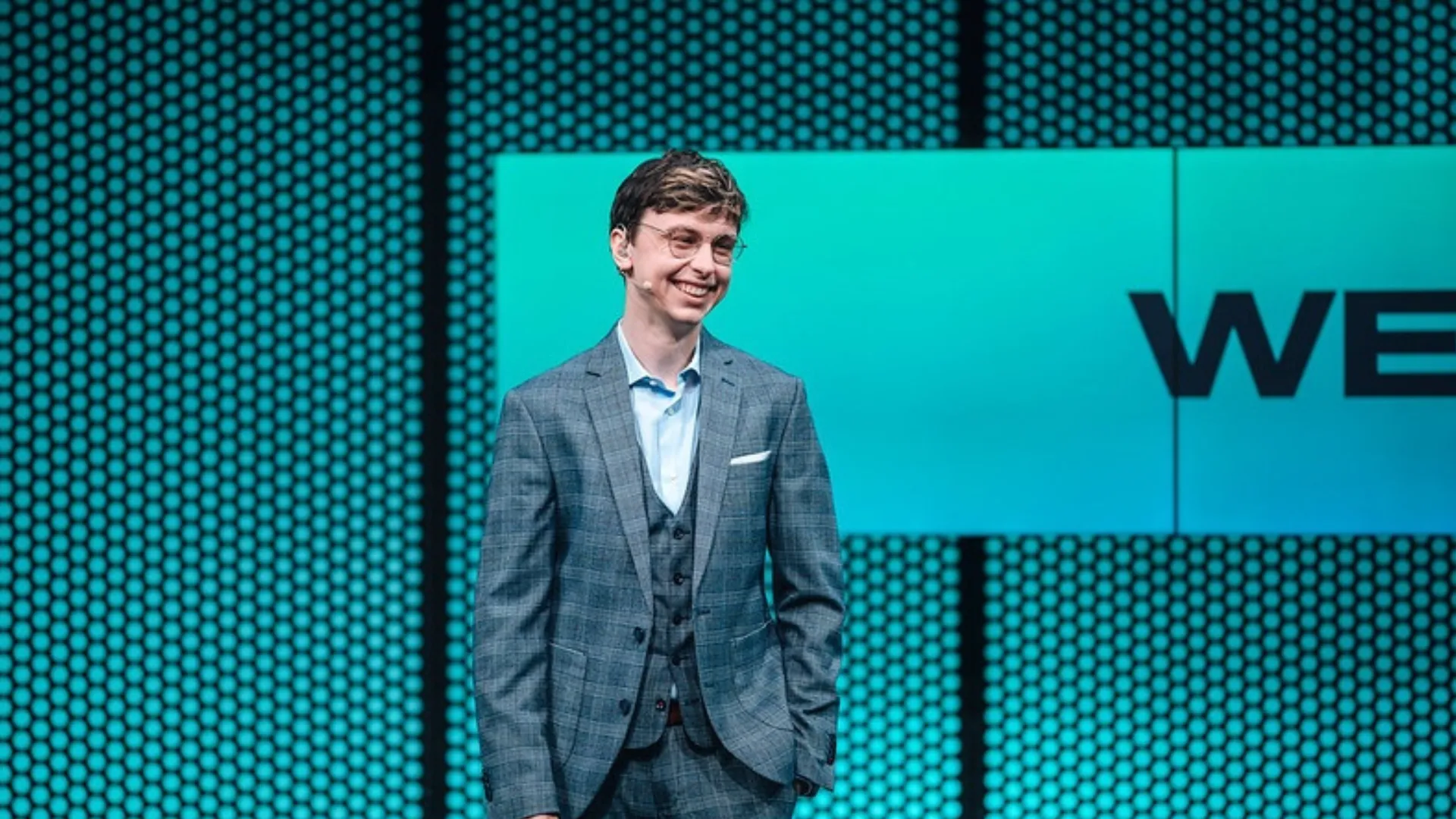 Caedrel smiles in a grey checked three-piece suit on a teal-lit stage with a large screen behind him.
