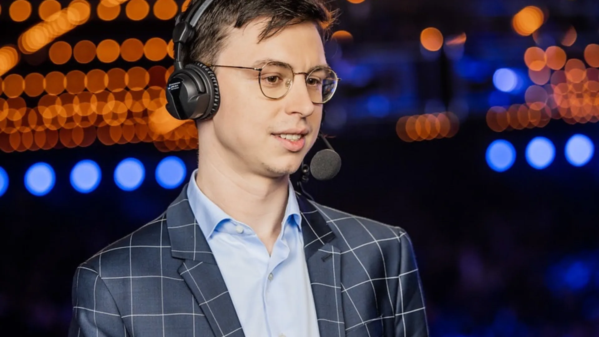 Caedrel in glasses and a checked blazer speaks into a headset mic with warm and blue arena lights blurred behind him.
