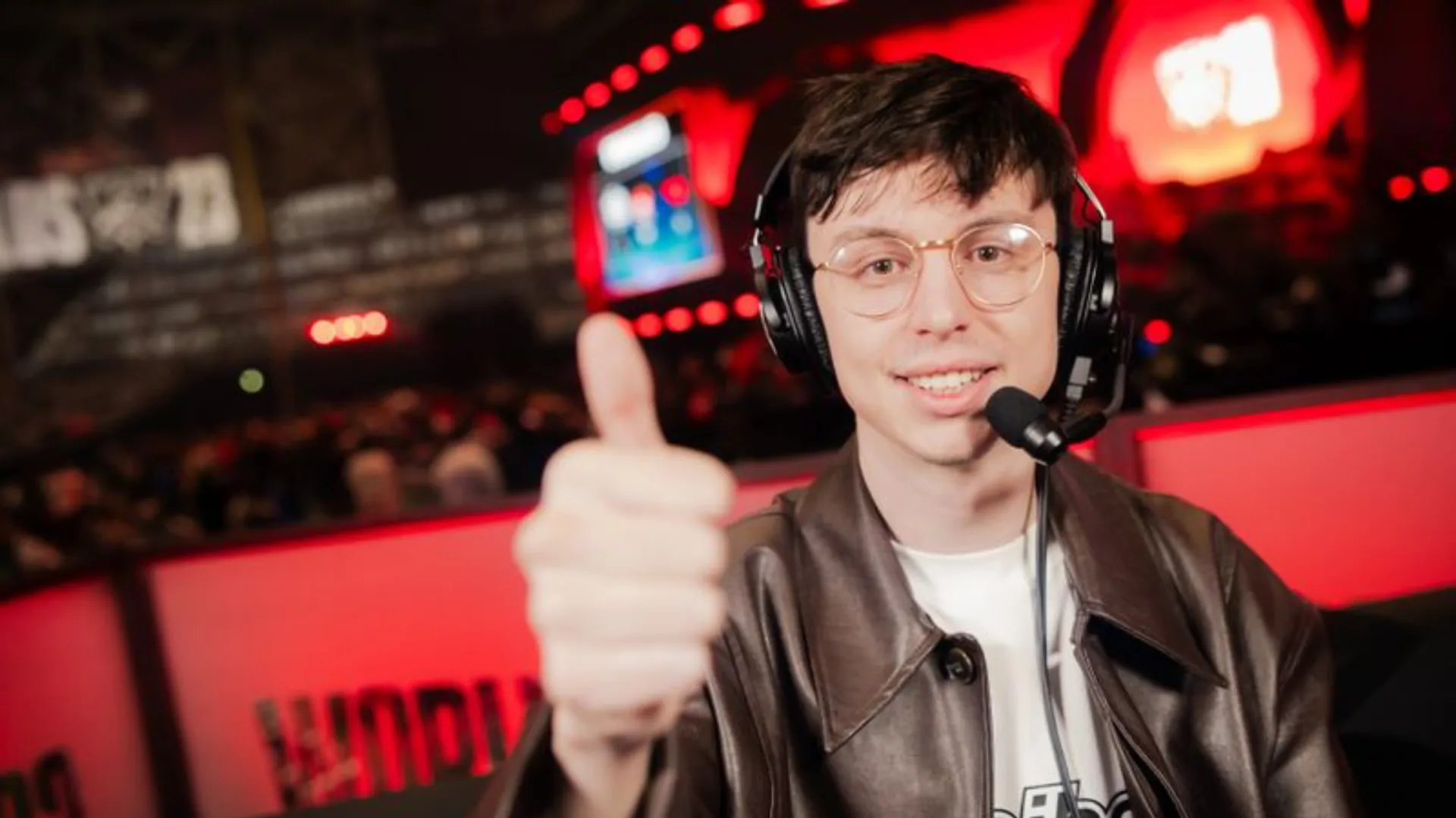 Caedrel in a leather jacket and headset gives a thumbs up to the camera at a red-lit League of Legends arena.