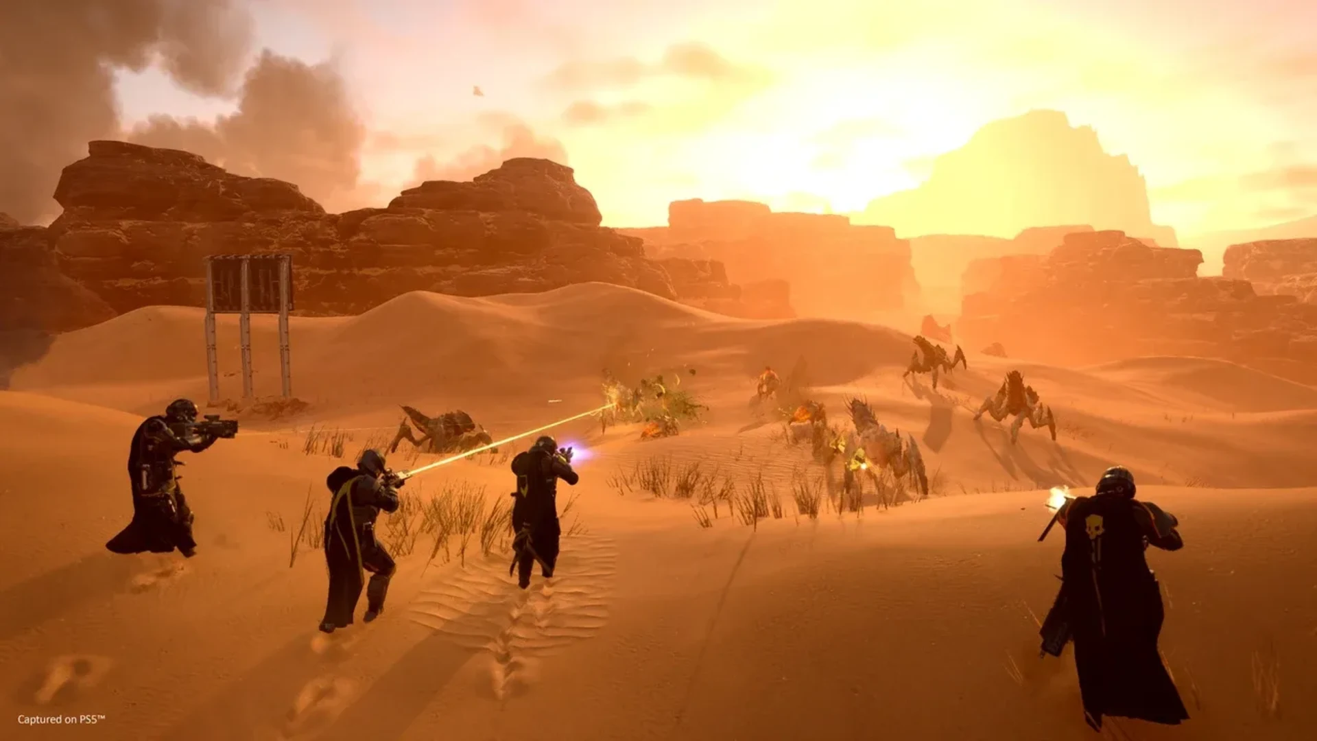 Four Helldivers fire at approaching Terminids across an orange desert battlefield.