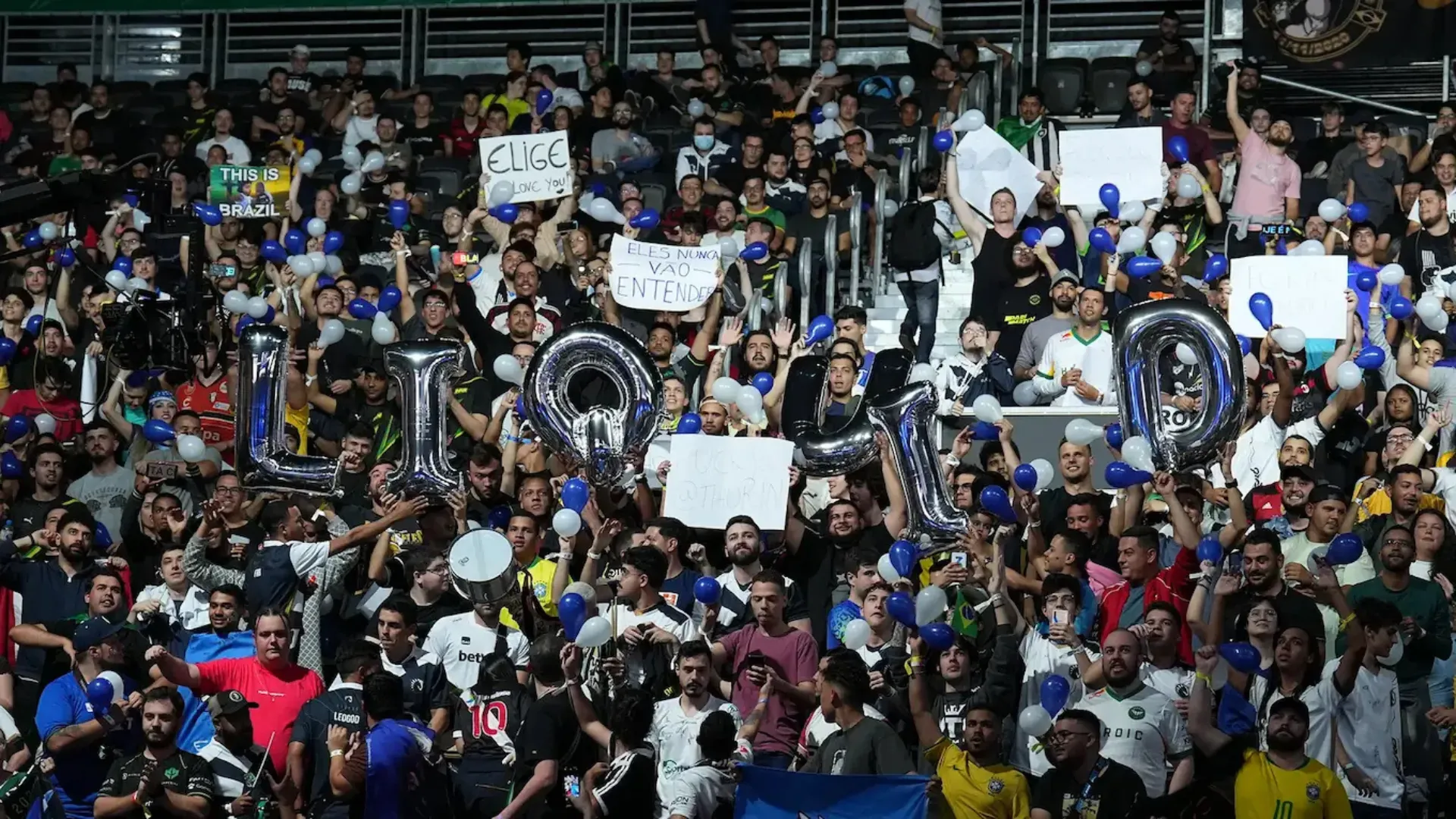 A packed crowd holds silver balloon letters spelling LIQUID and handmade signs at a live esports event.