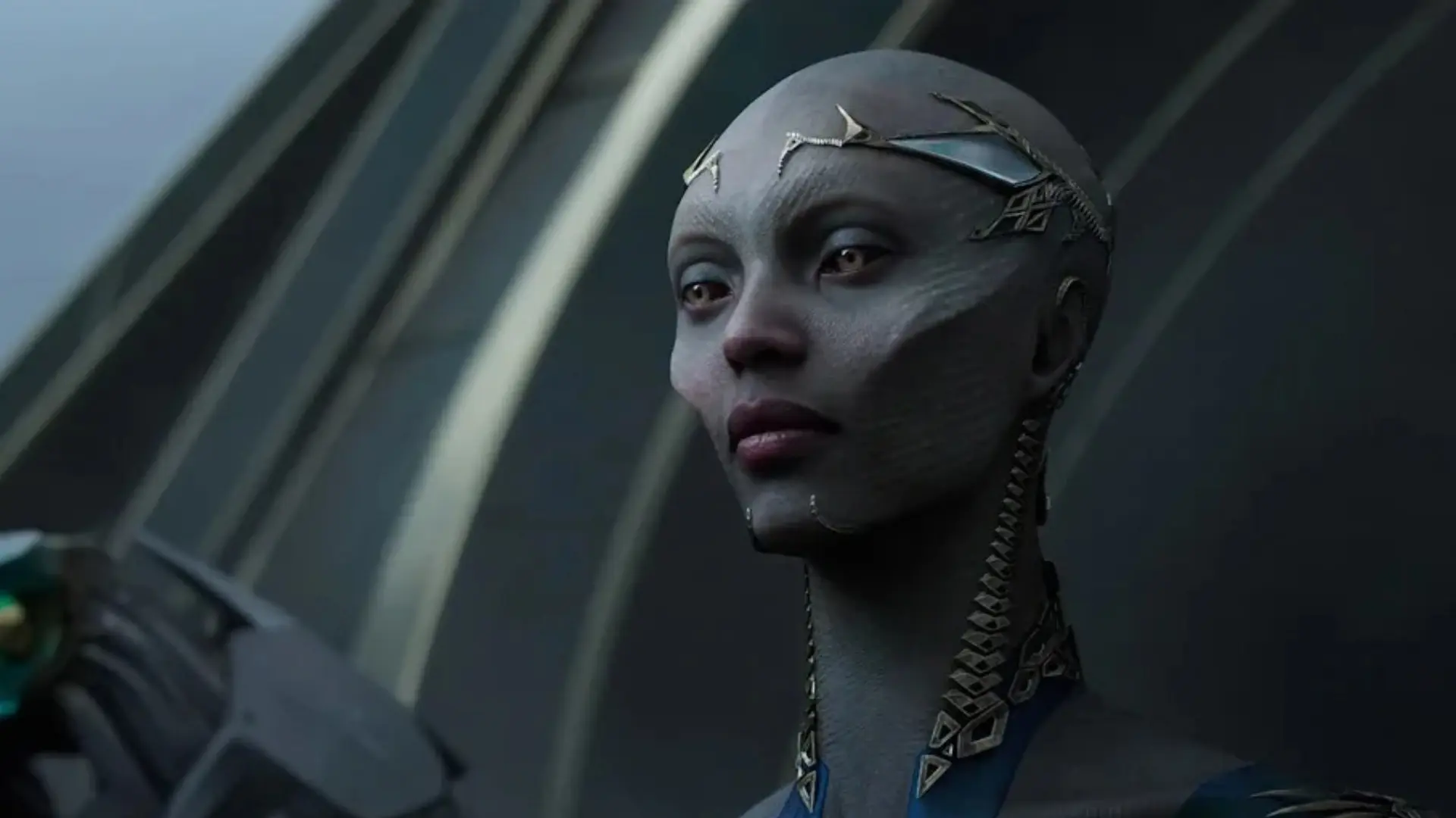A gray-skinned woman with metallic facial adornments appears in a close-up from EXODUS