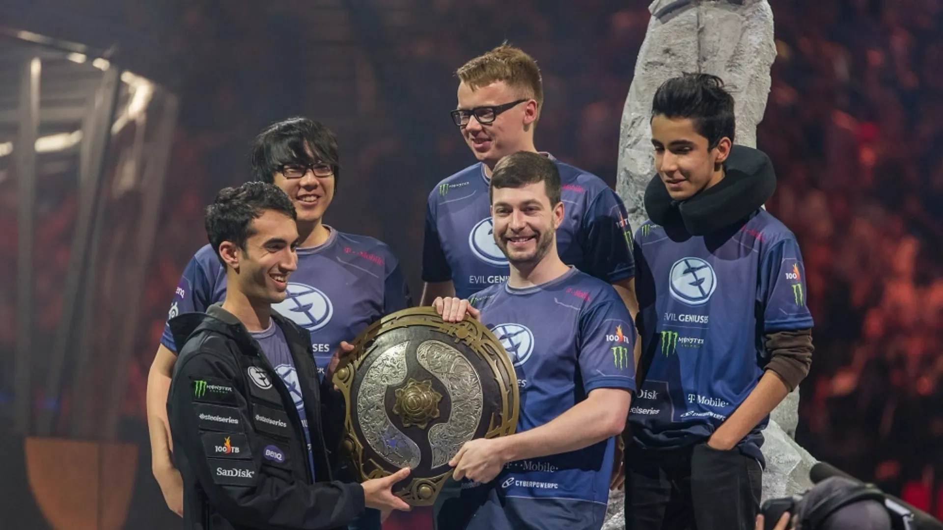 Five Evil Geniuses Dota 2 players in blue jerseys holding the Aegis of Champions on stage at The International 2015.