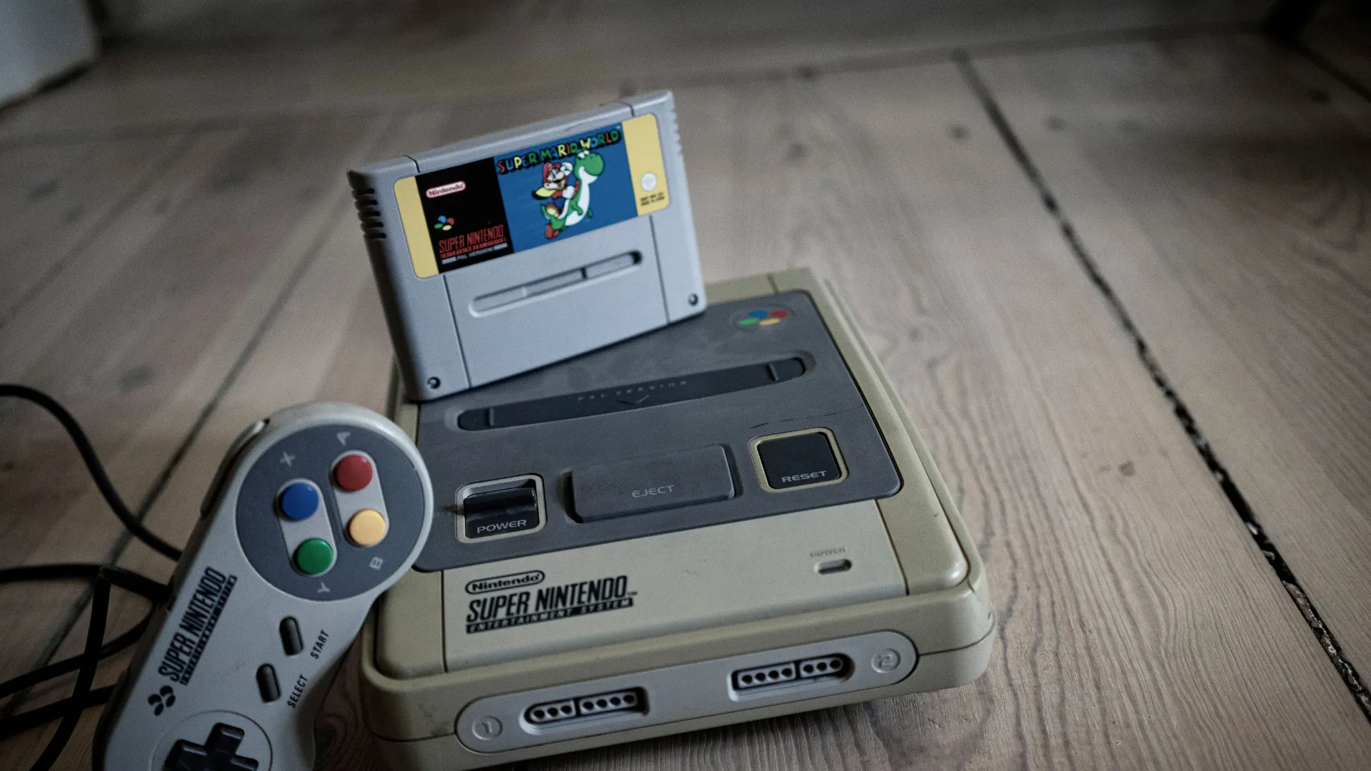 A Super Nintendo console on a wooden floor with a Super Mario World cartridge inserted and a SNES controller beside it.