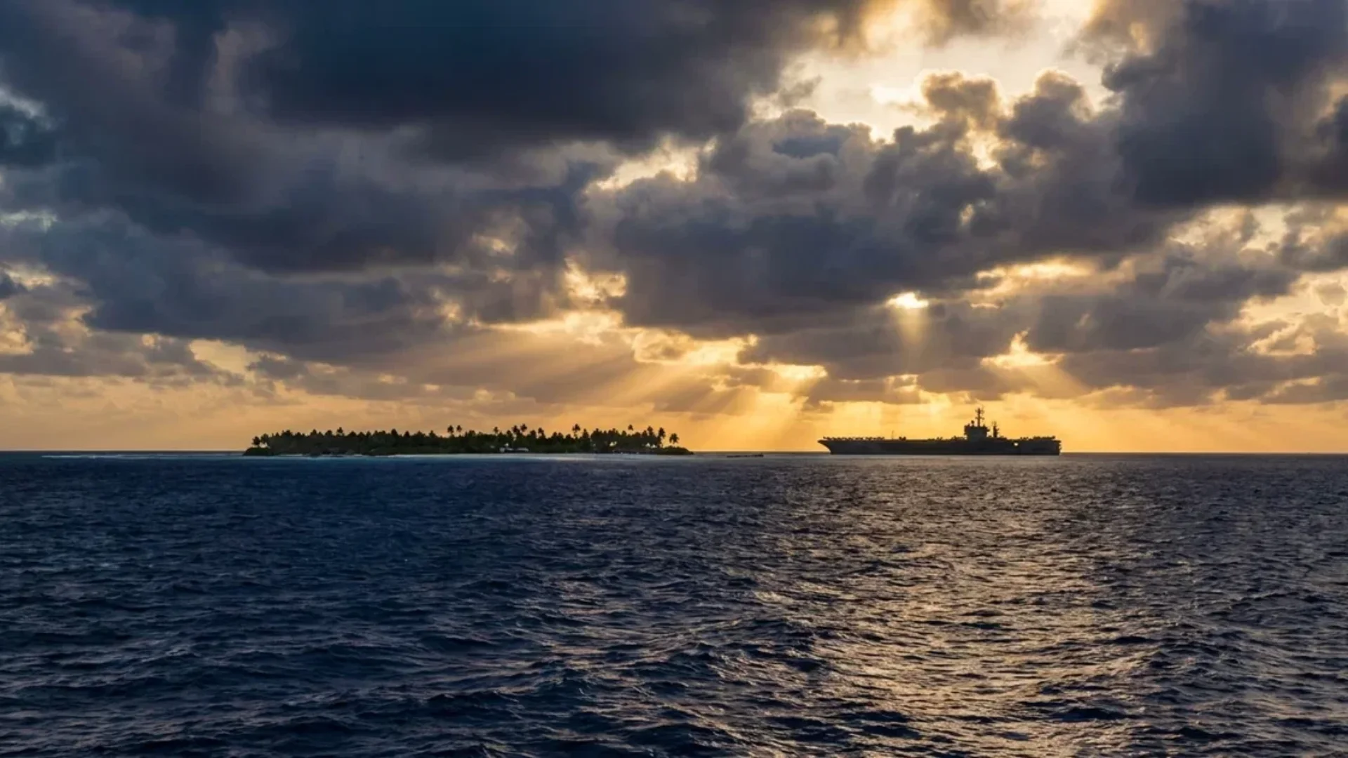 Aircraft carrier sails near a tropical island beneath dark clouds and rays of sunlight.
