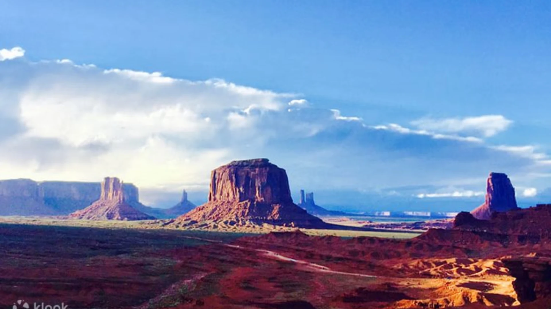 Red sandstone buttes and mesas stretch across Monument Valley beneath a bright blue sky and large clouds.