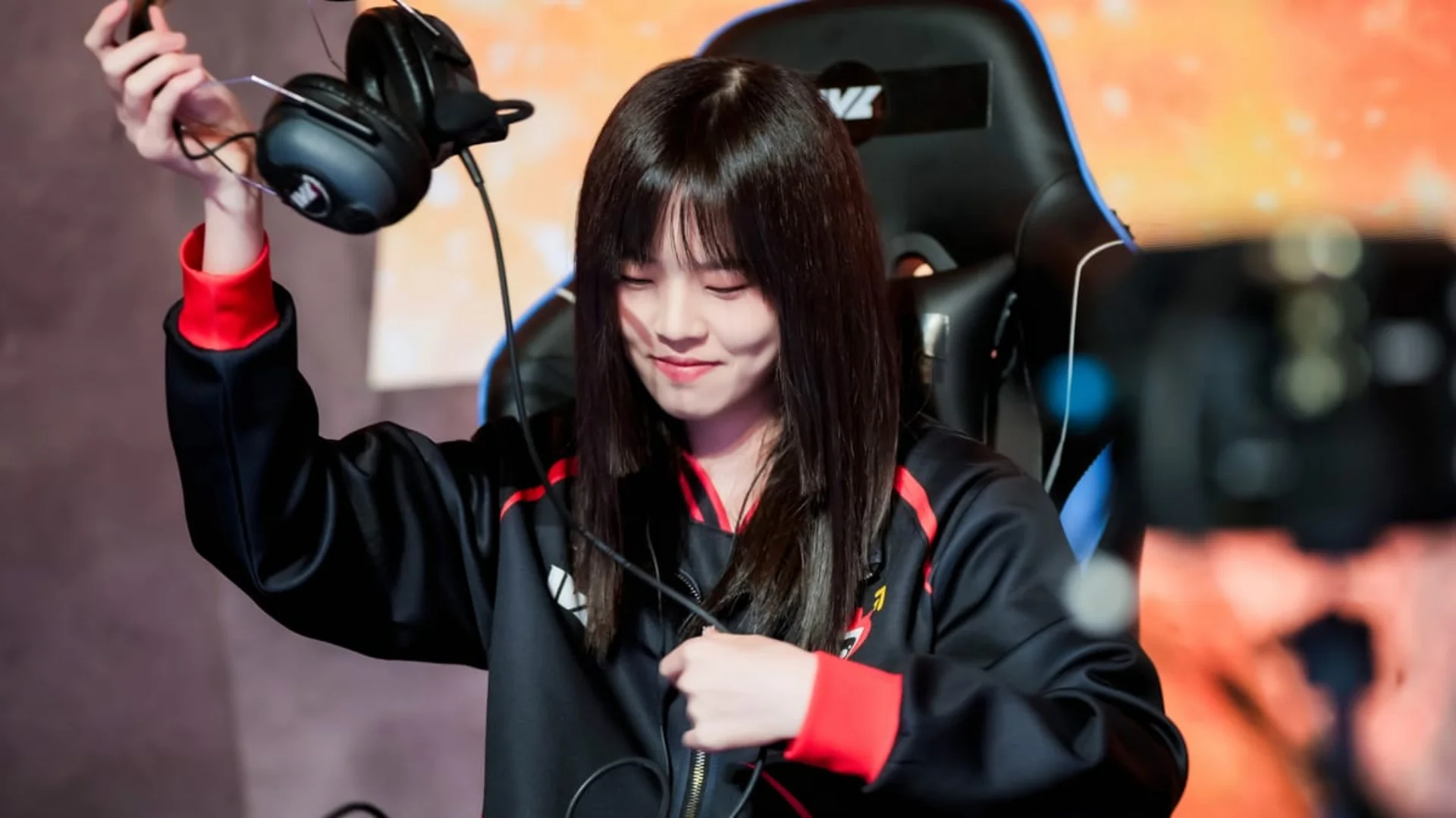 A female esports player in a black and red team jacket raises her headset while seated at a gaming station.