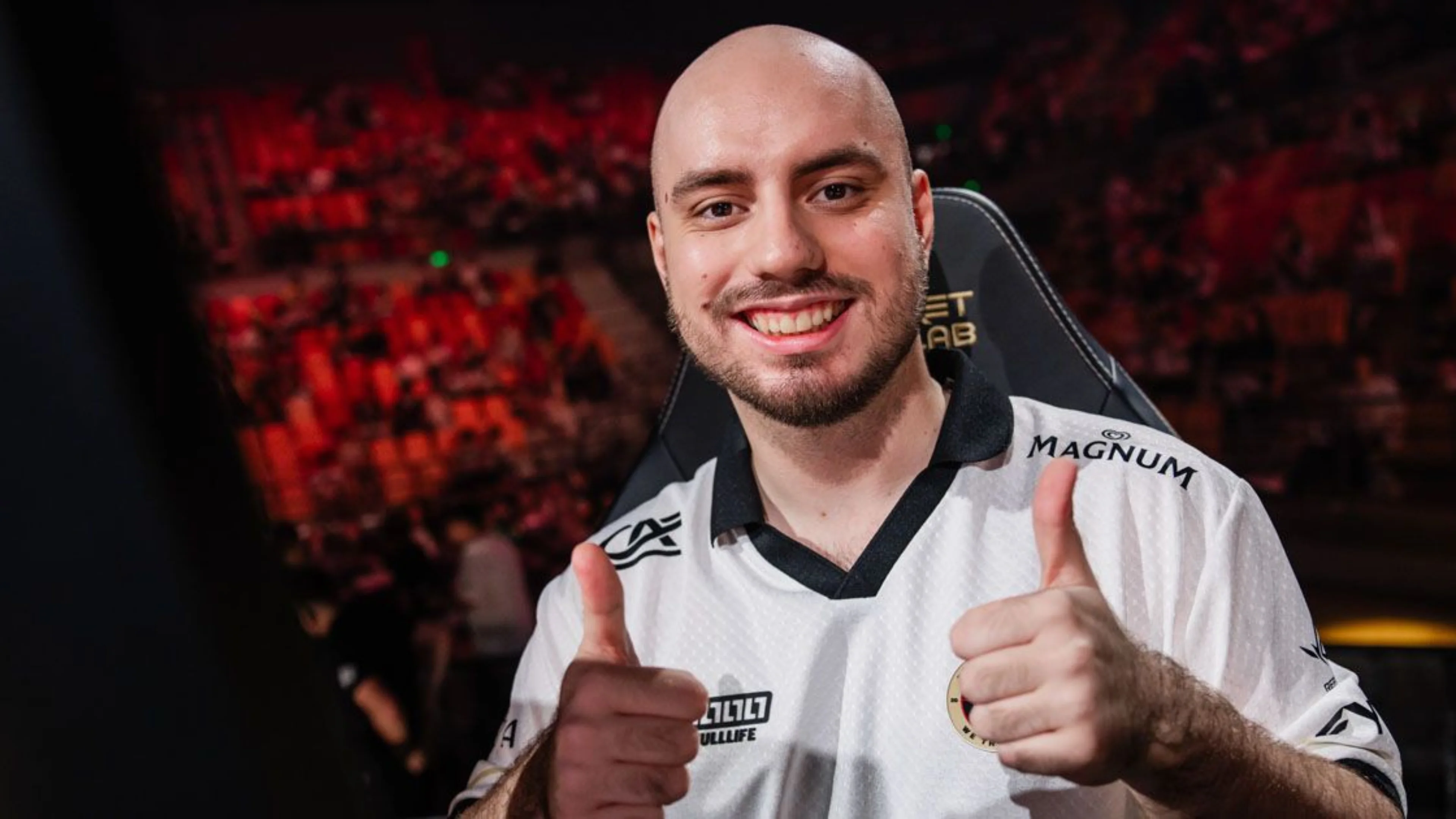 A smiling VALORANT player in a white Team Vitality jersey gives two thumbs up while seated at a tournament gaming station.