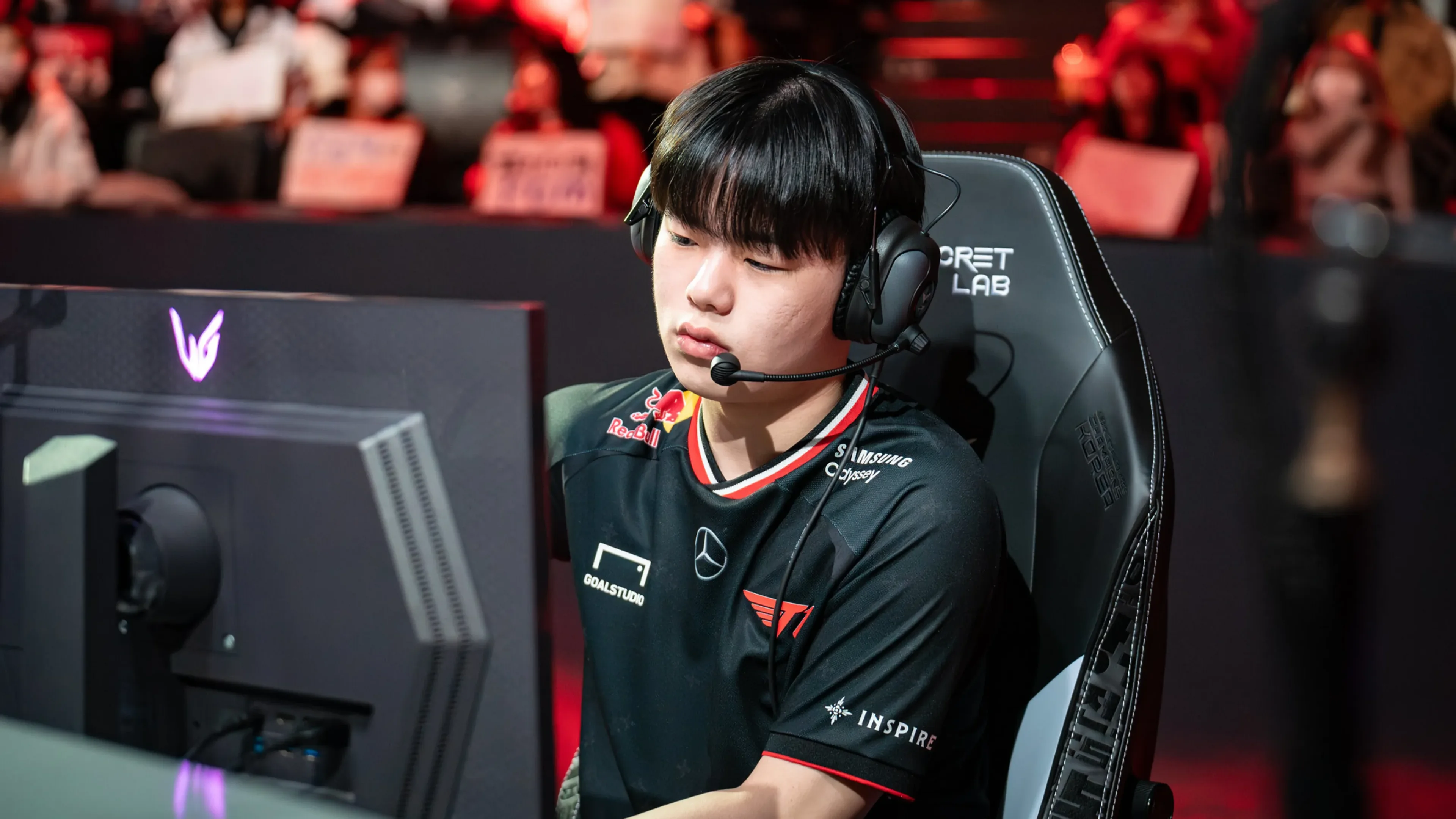A League of Legends player wearing a black T1 jersey and gaming headset concentrates on a monitor during an esports match.