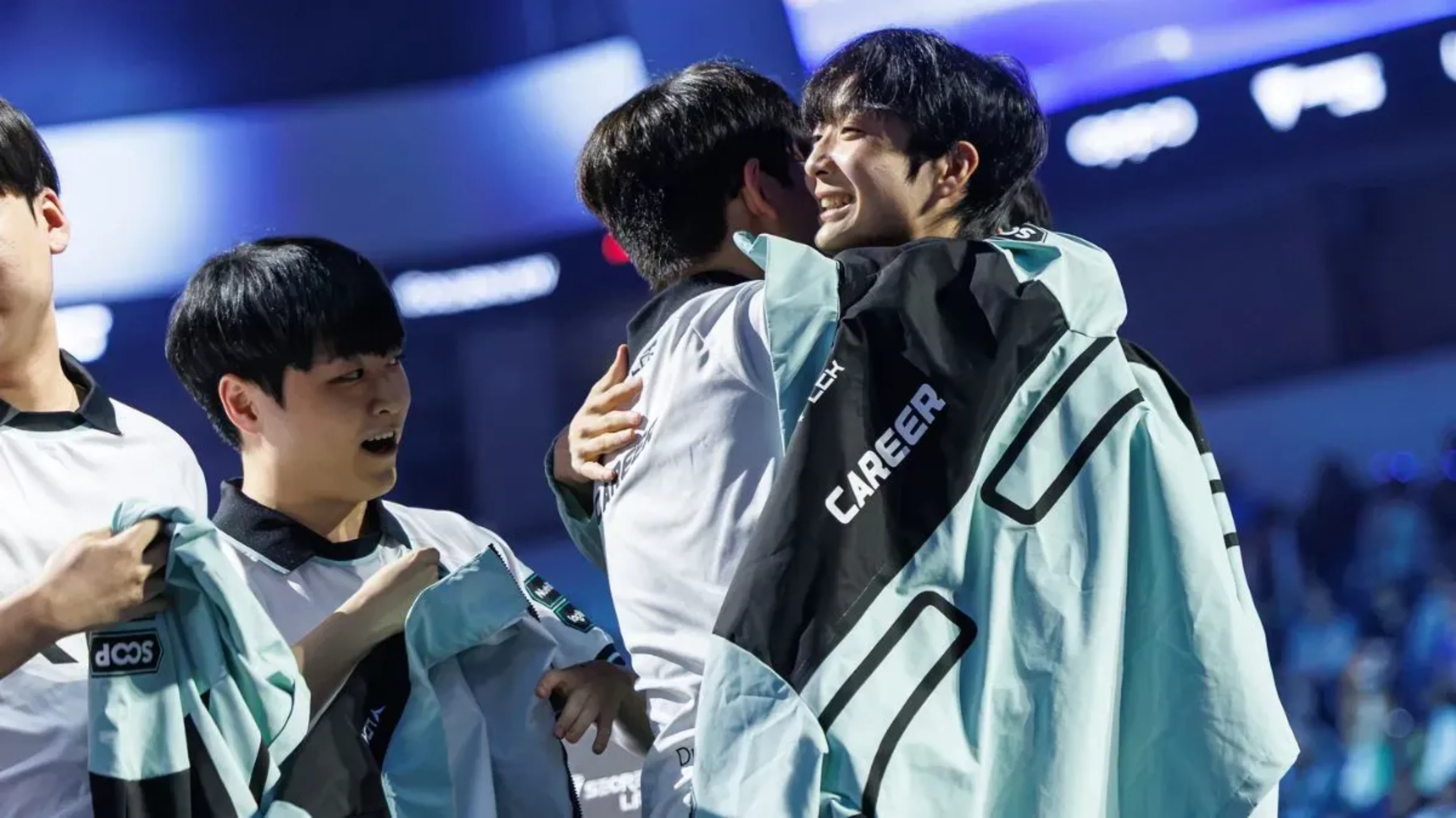 League of Legends esports players in white and light blue team jackets embrace and celebrate together on stage.