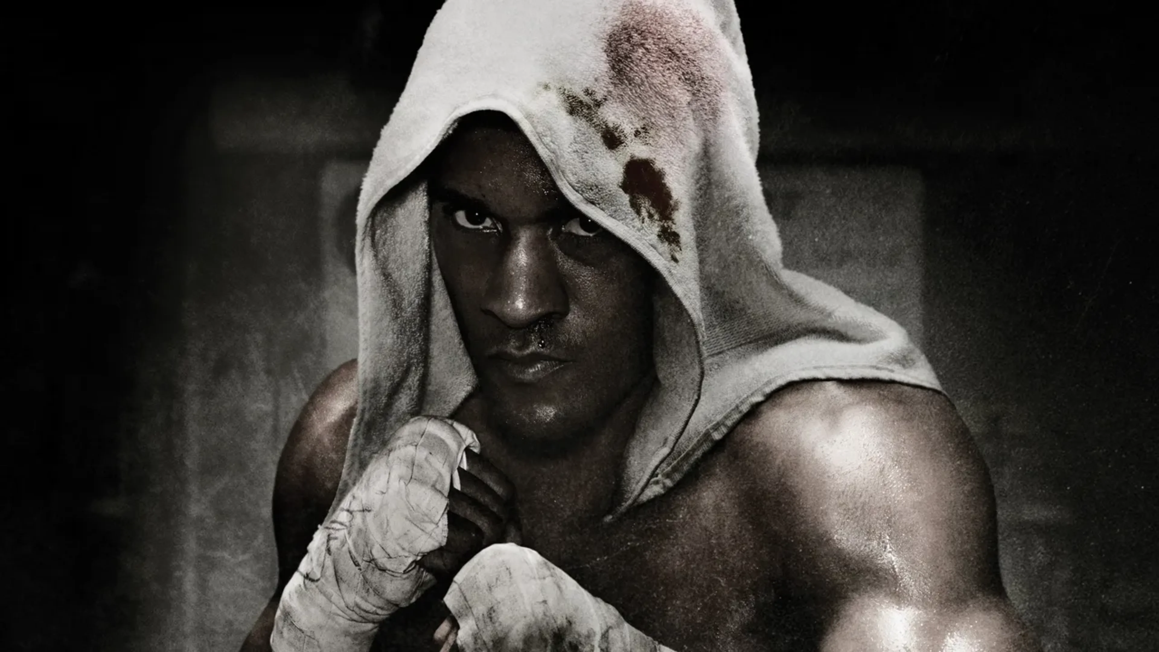 Black-and-white portrait of a muscular boxer wearing a stained white hood and hand wraps while holding a fighting stance.