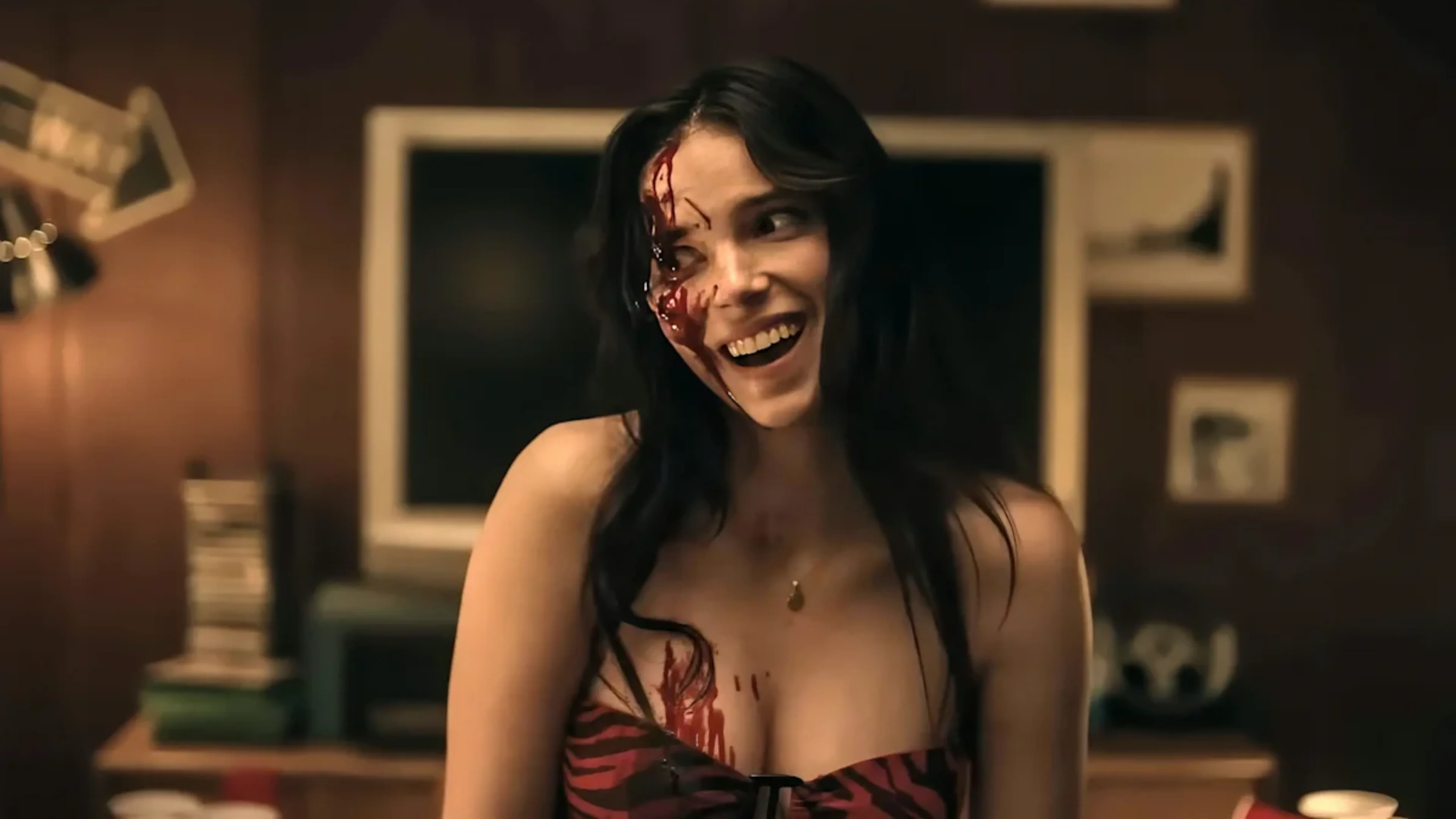 Woman with long dark hair smiles with blood across one side of her face in a dimly lit room in Obsession.
