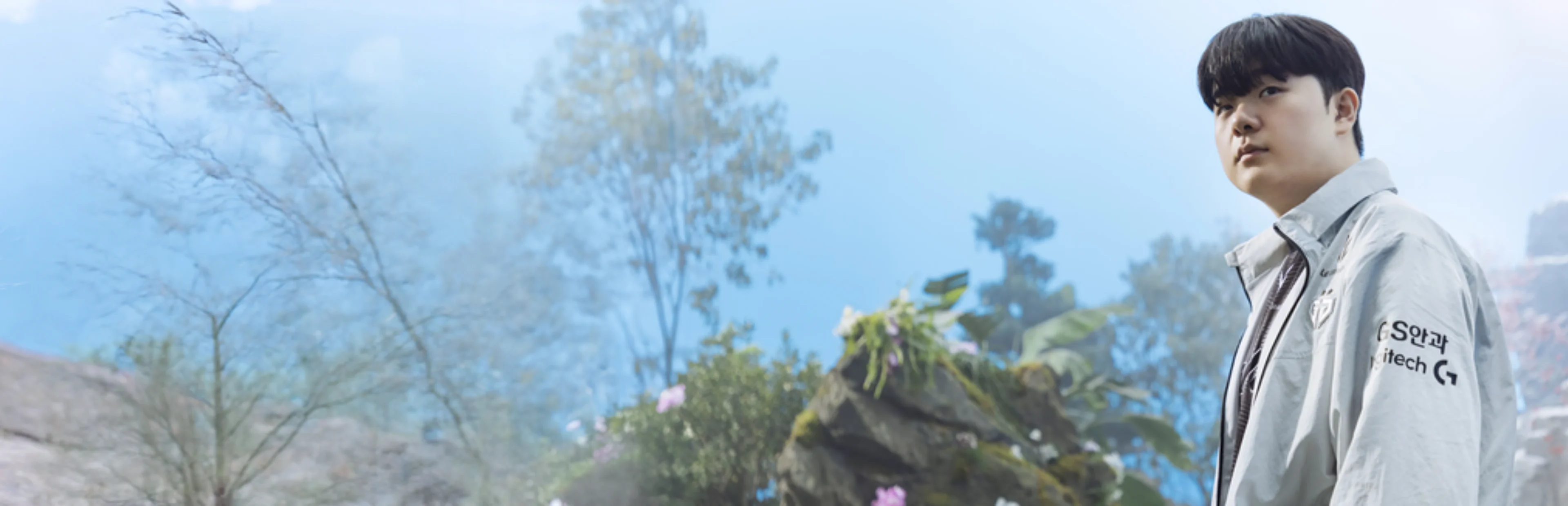 Gen.G jungler Canyon wears a white team jacket with sponsor logos while standing against a softly blurred outdoor backdrop.