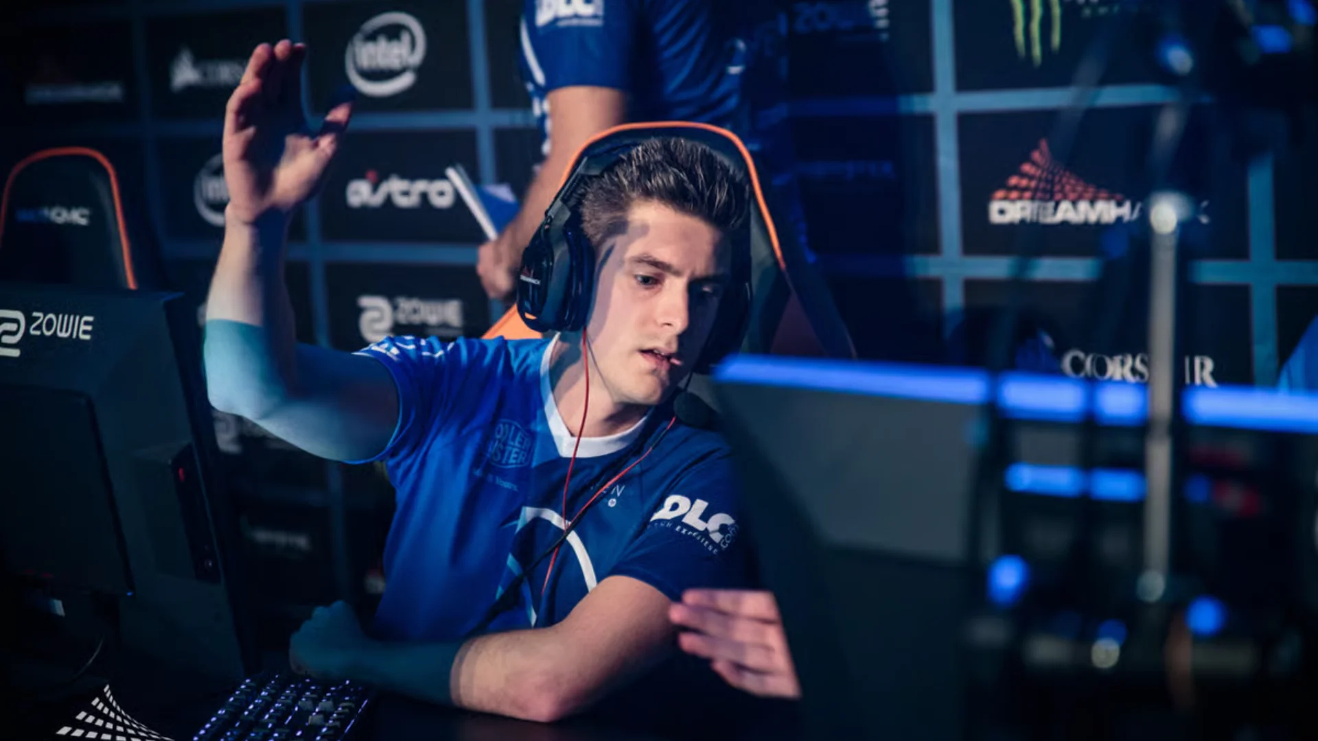 Esports coach wearing a blue team jersey and headset raises one hand while seated at a tournament PC.