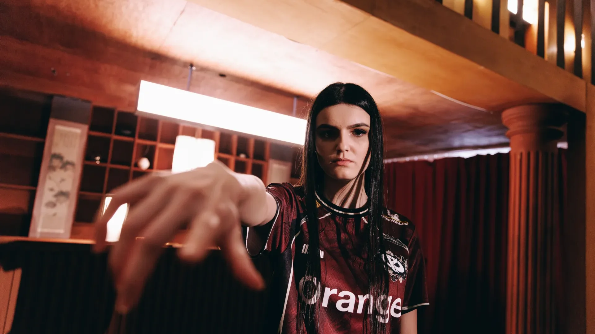 Woman wearing a Gentle Mates esports jersey reaches toward the camera inside a warmly lit room.