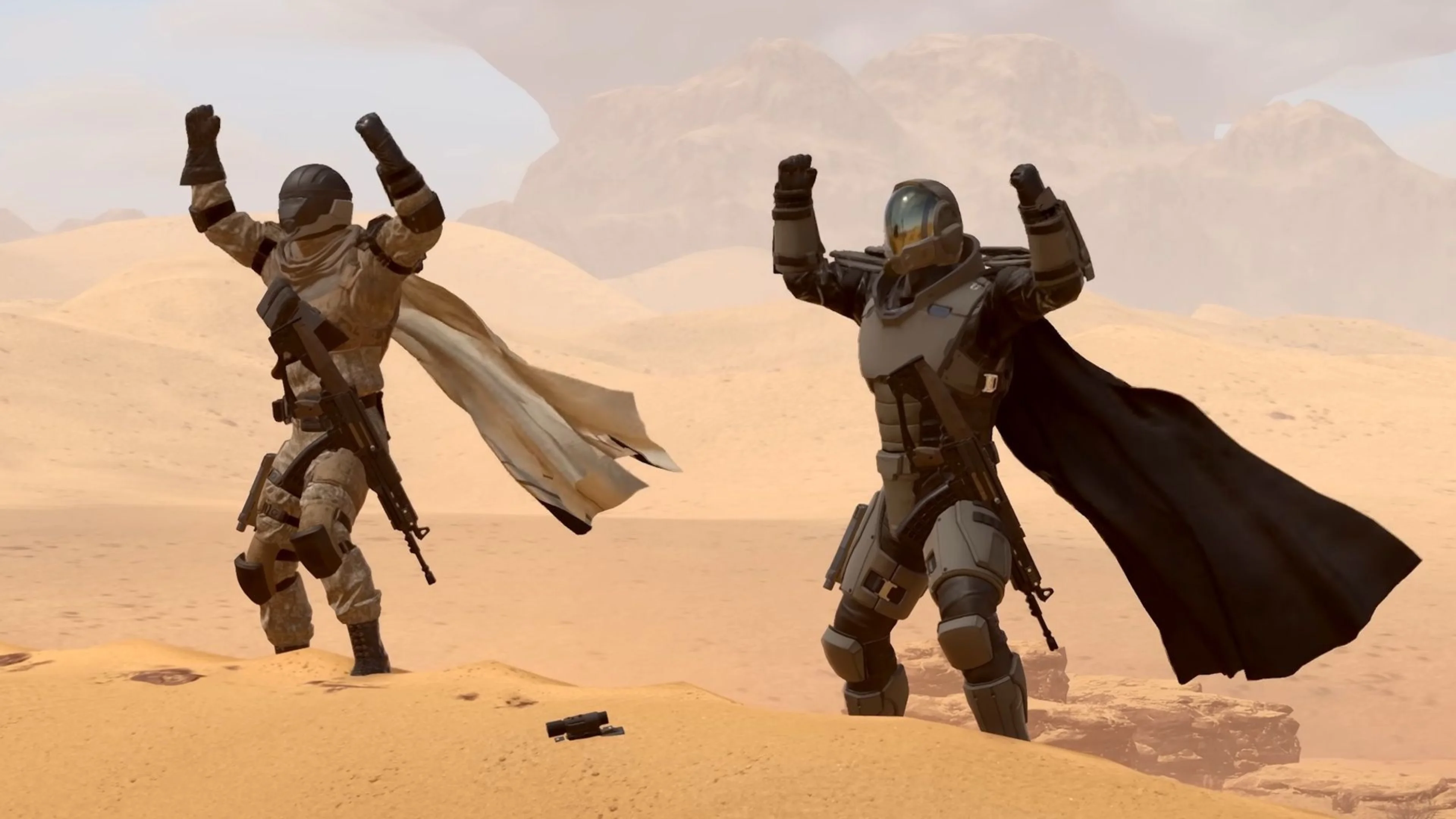Two armored Helldivers wearing flowing capes raise their fists while standing on a sandy desert planet in Helldivers 2.