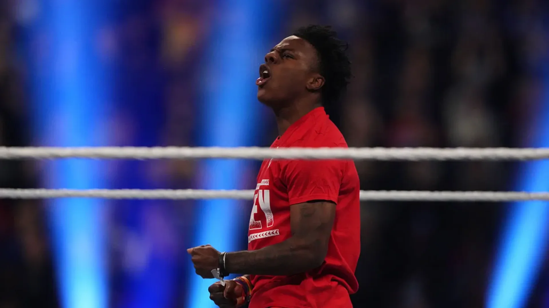 A streamer in a red shirt celebrates inside a wrestling ring with blue arena lights in the background.