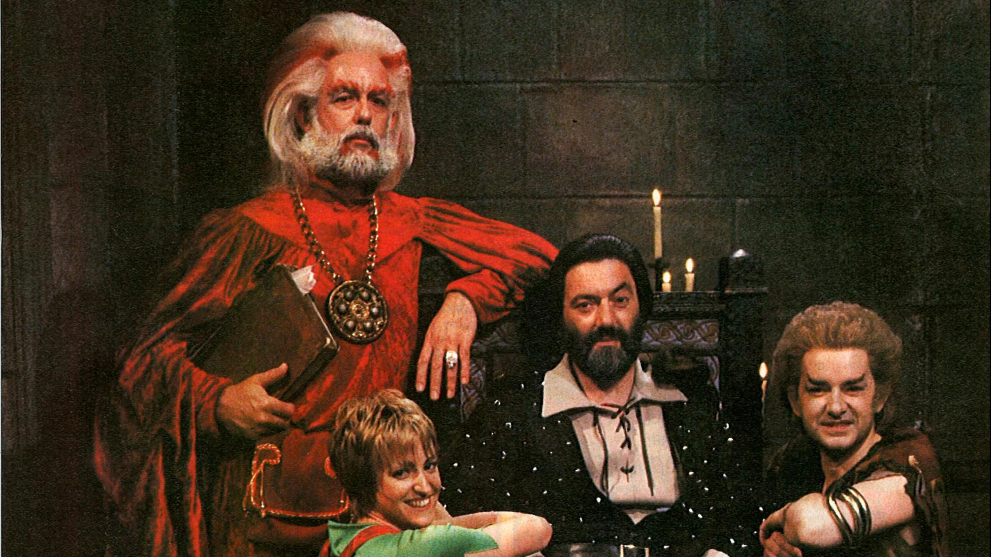 Four performers from the original Knightmare television series pose in medieval fantasy costumes inside a candlelit castle setting.