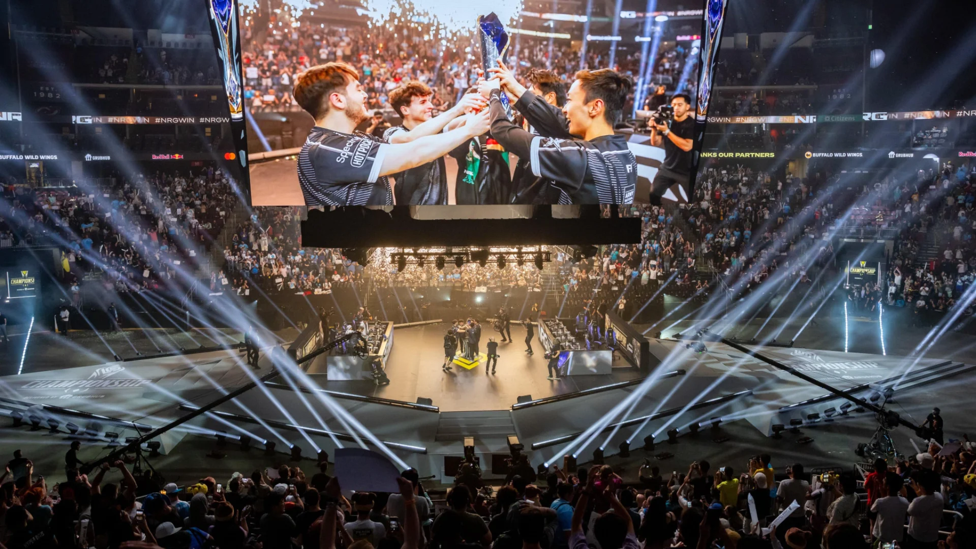 A packed League of Legends esports arena with a team lifting a trophy on a giant overhead screen.