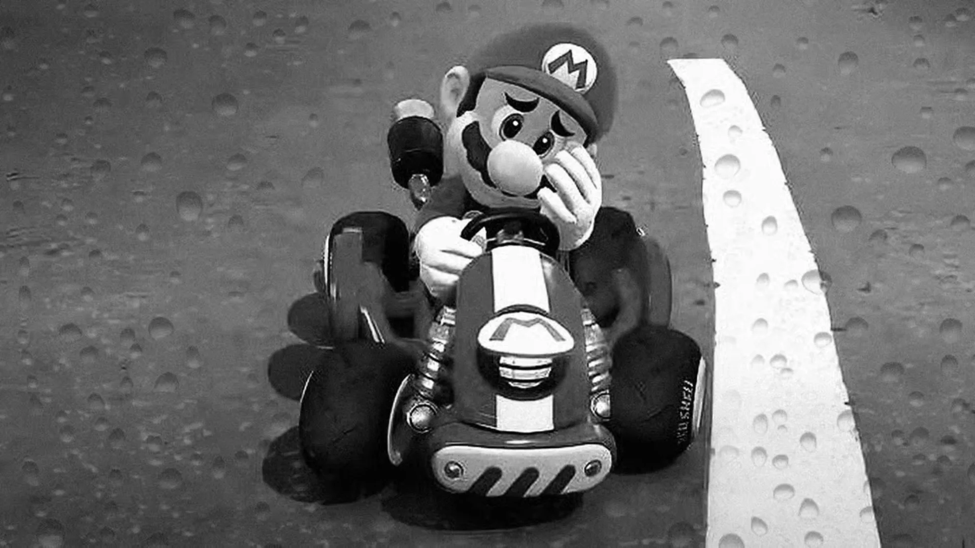 Mario sits in a kart with one hand against his face in a black-and-white Mario Kart image.