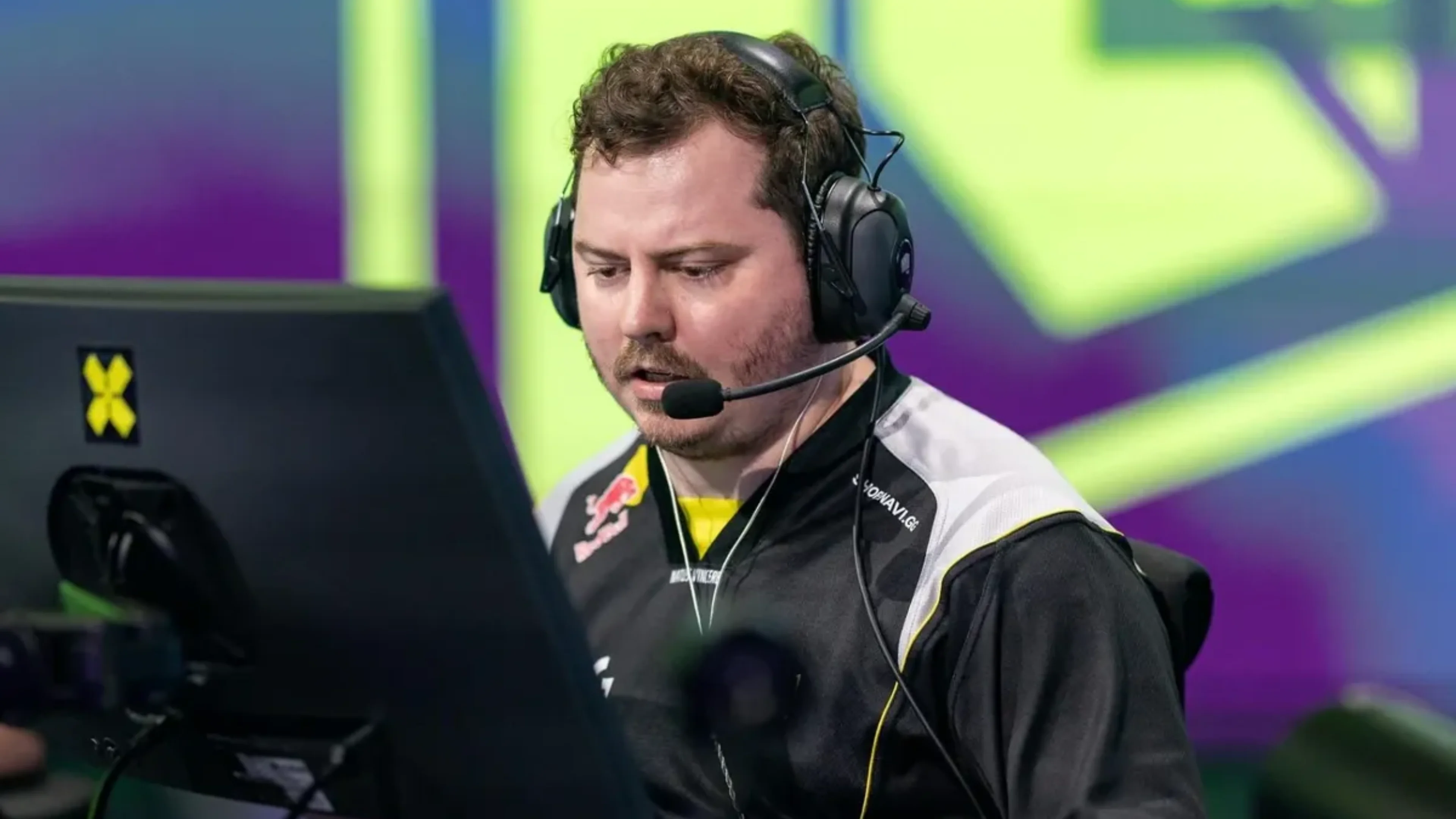 A NAVI VALORANT team member wearing a black-and-white jersey and gaming headset looks at a monitor during an esports event.