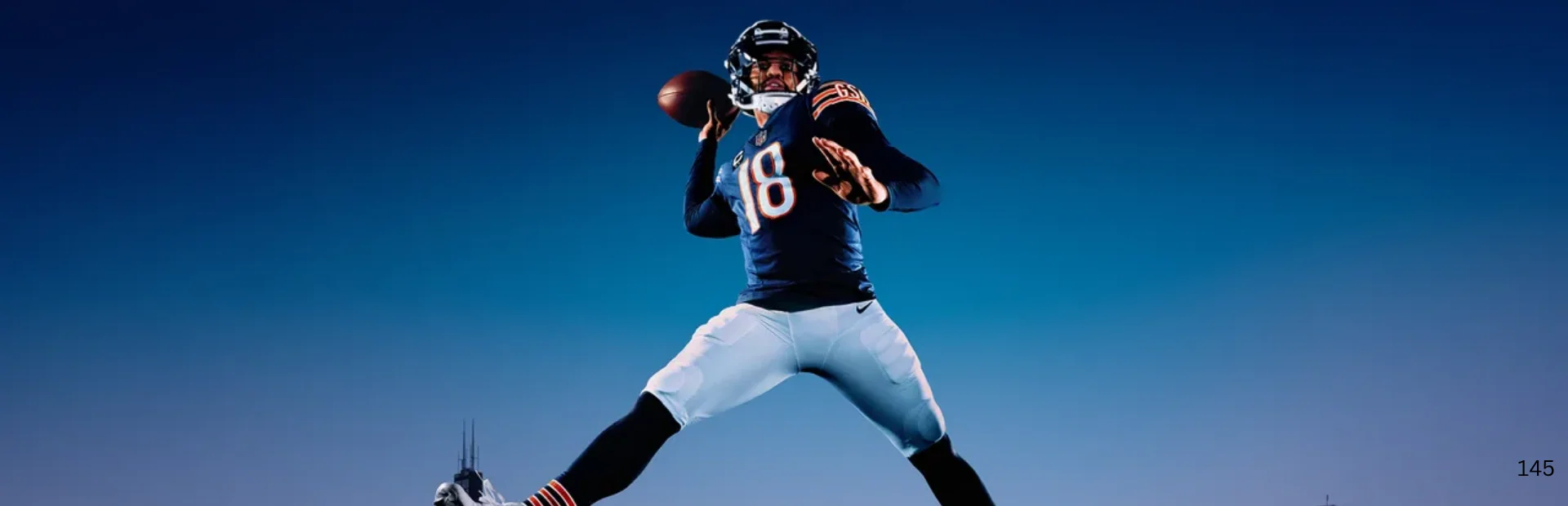 A football player in a navy jersey holds the ball in a throwing stance against a blue sky.