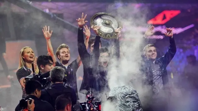 OG players celebrating a Dota 2 championship victory while lifting the Aegis trophy on stage.