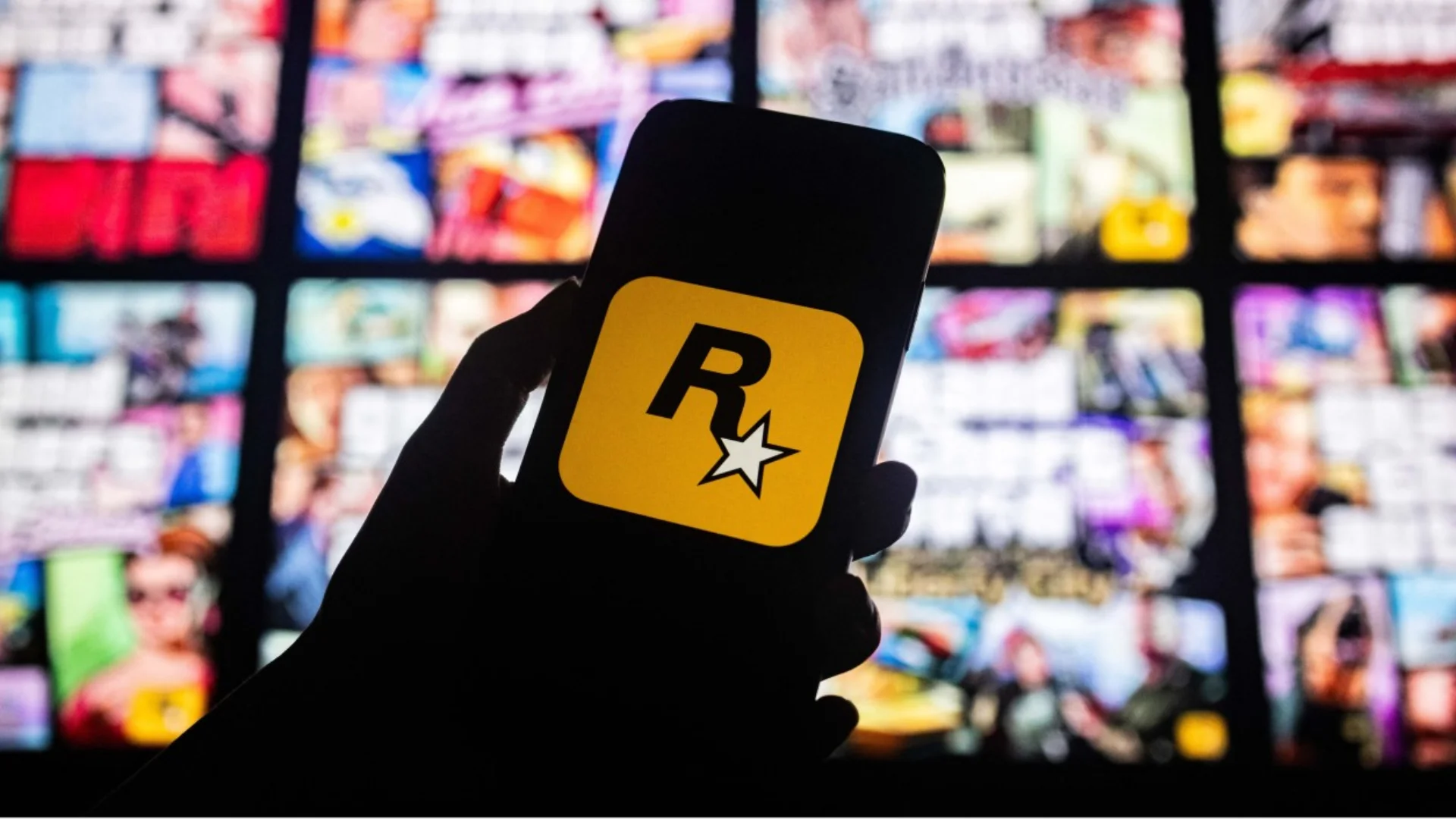 A silhouetted hand holds a smartphone displaying the Rockstar Games logo in front of blurred screens.