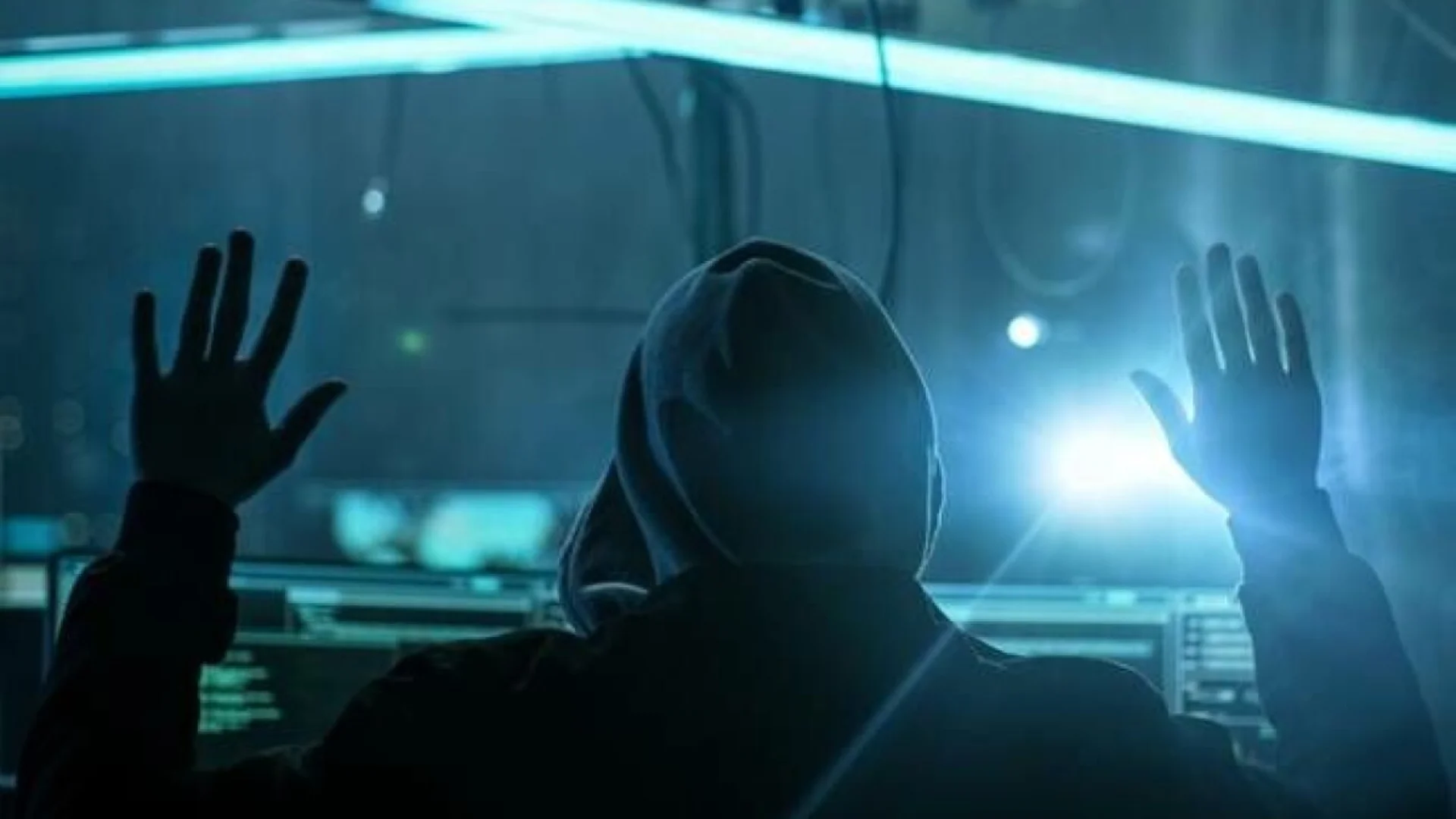 A hooded figure raises both hands in front of computer screens and bright blue lighting.
