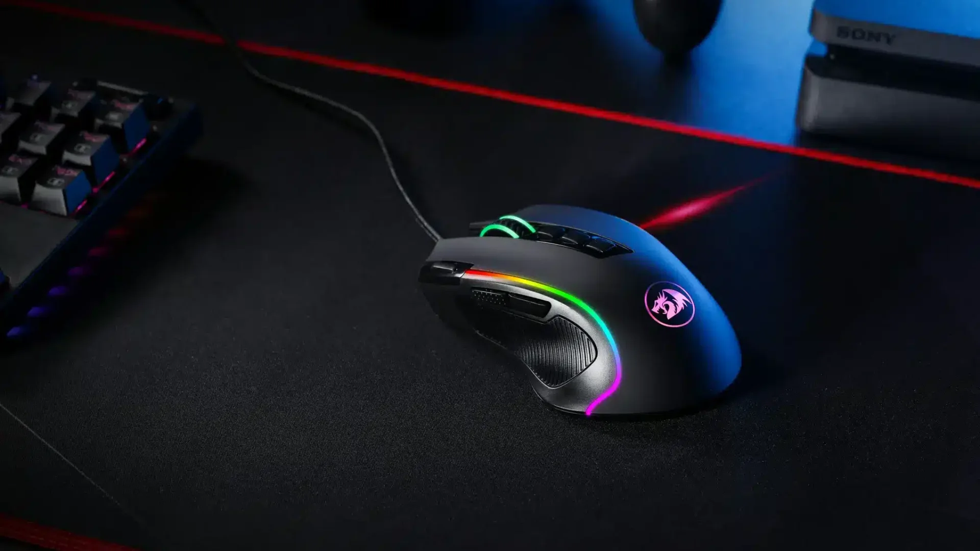A wired RGB gaming mouse rests on a large black mouse pad beside a mechanical keyboard.