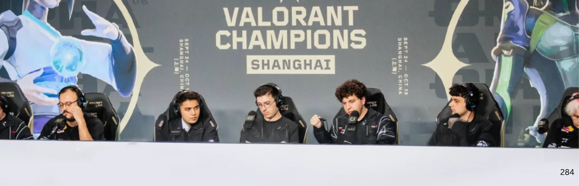 VALORANT esports players sit at tournament PCs beneath VALORANT Champions Shanghai branding.