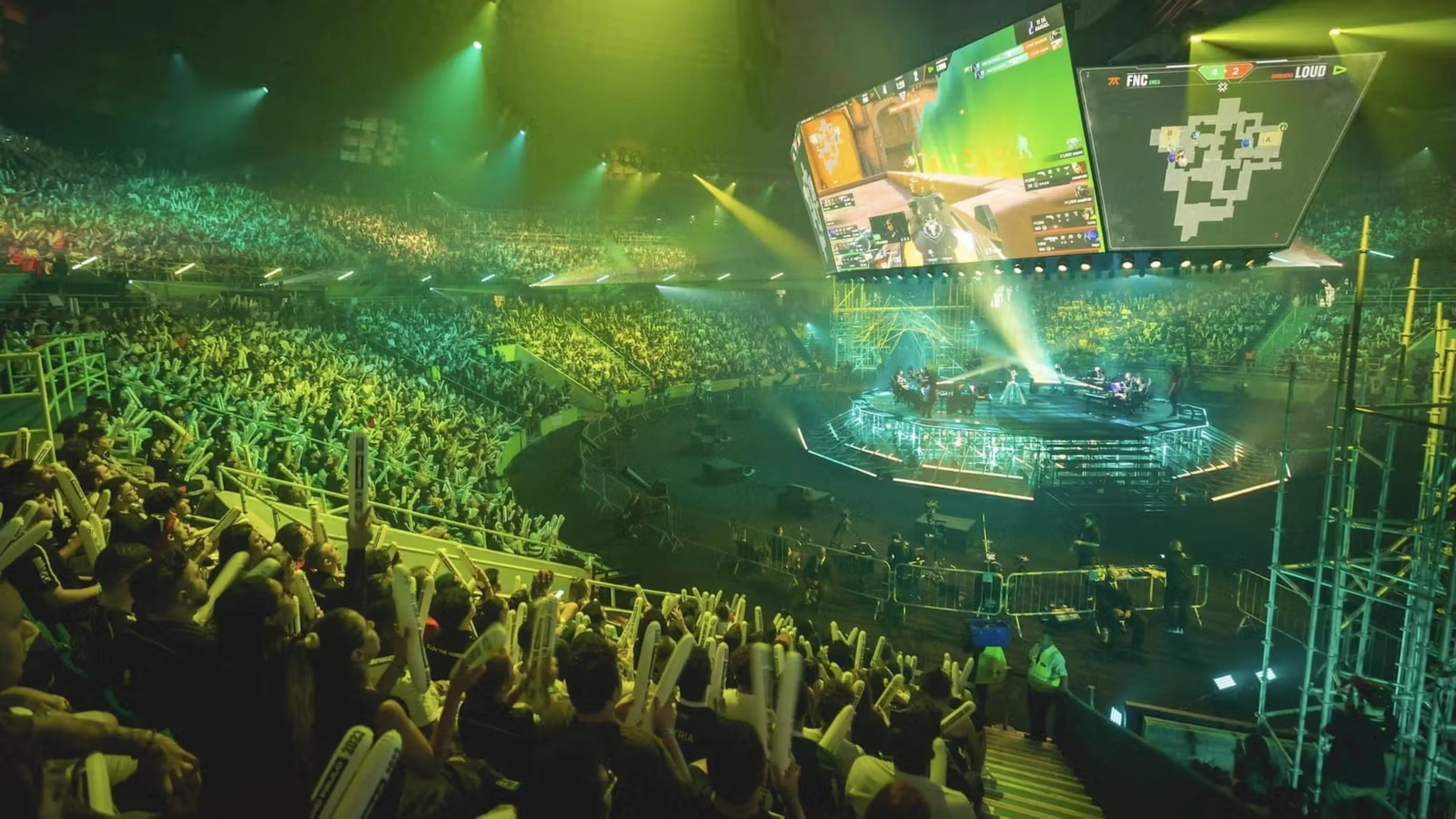 Packed VALORANT esports arena with fans watching a live match on a central stage under green spotlights.