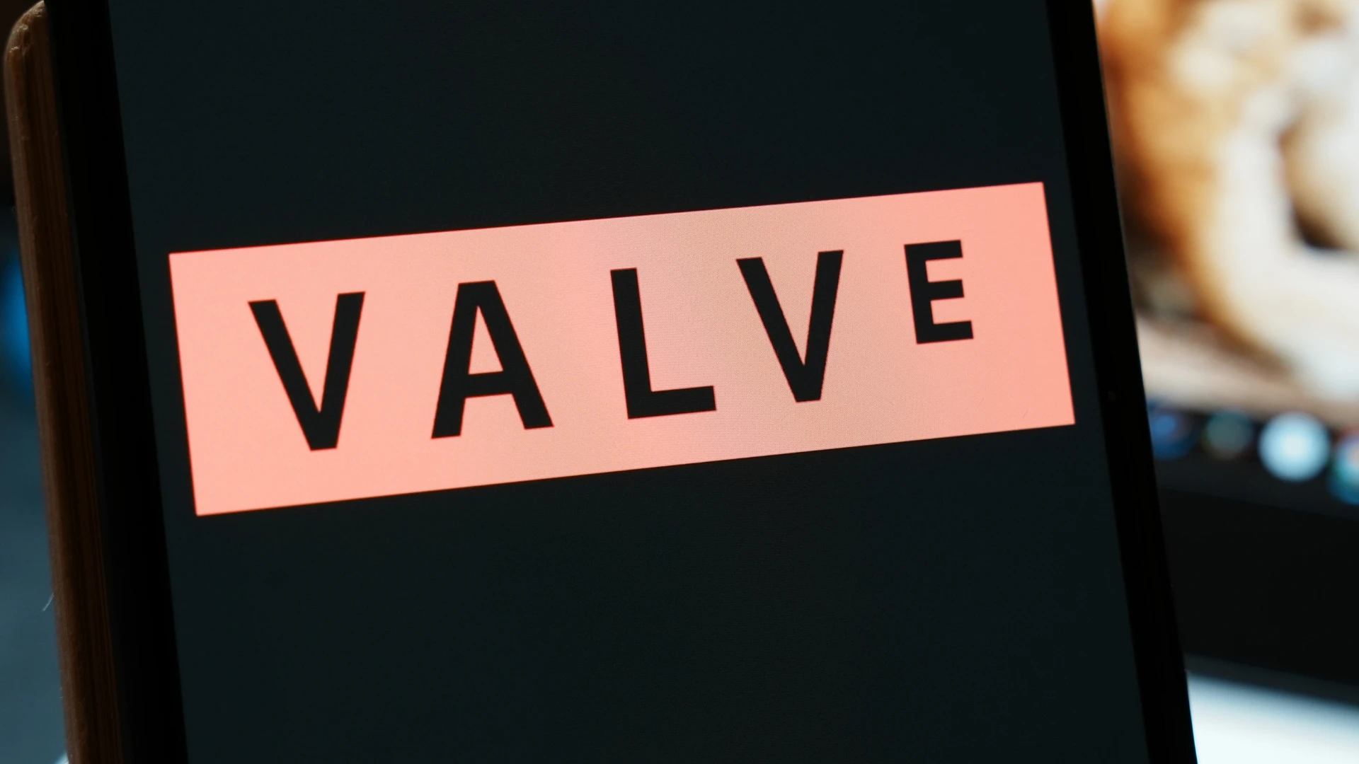 Valve logo displayed on a mobile device screen with a blurred gaming controller in the background.