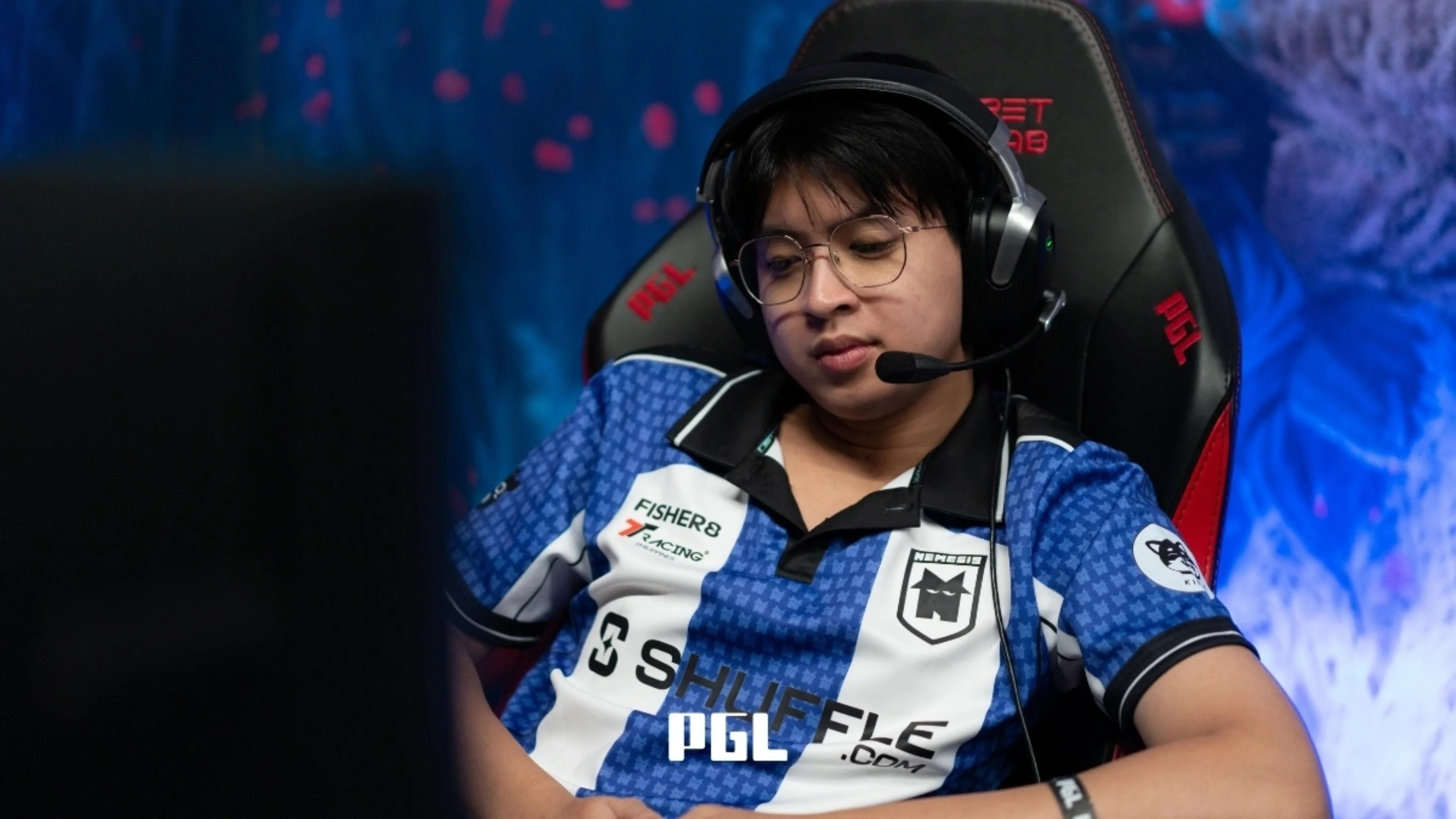 Professional Dota 2 player wearing glasses, a headset and a blue-and-white esports jersey while seated at a gaming station.