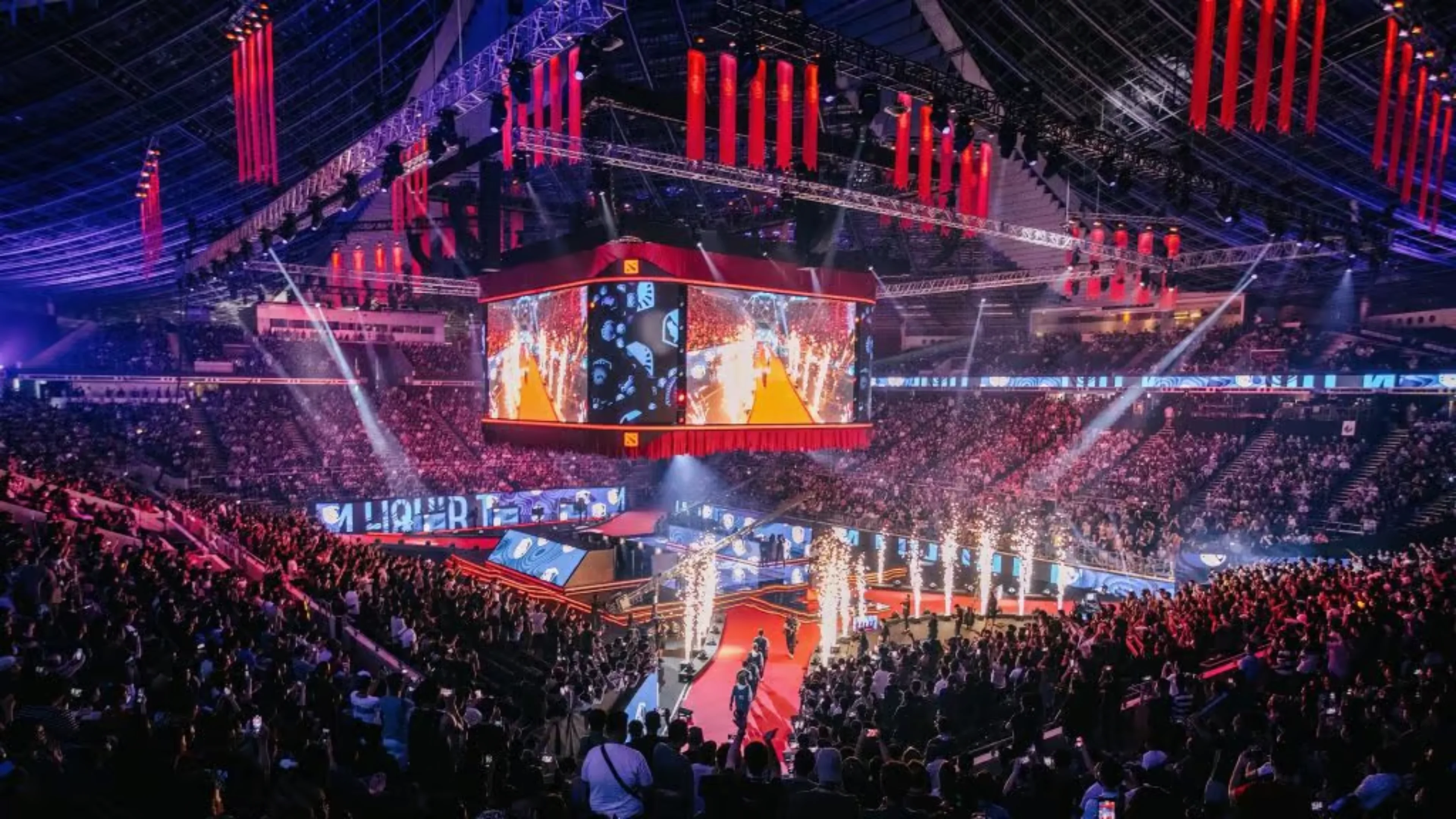 Packed Dota 2 tournament arena with overhead screens, red lighting and pyrotechnics during a live esports event.