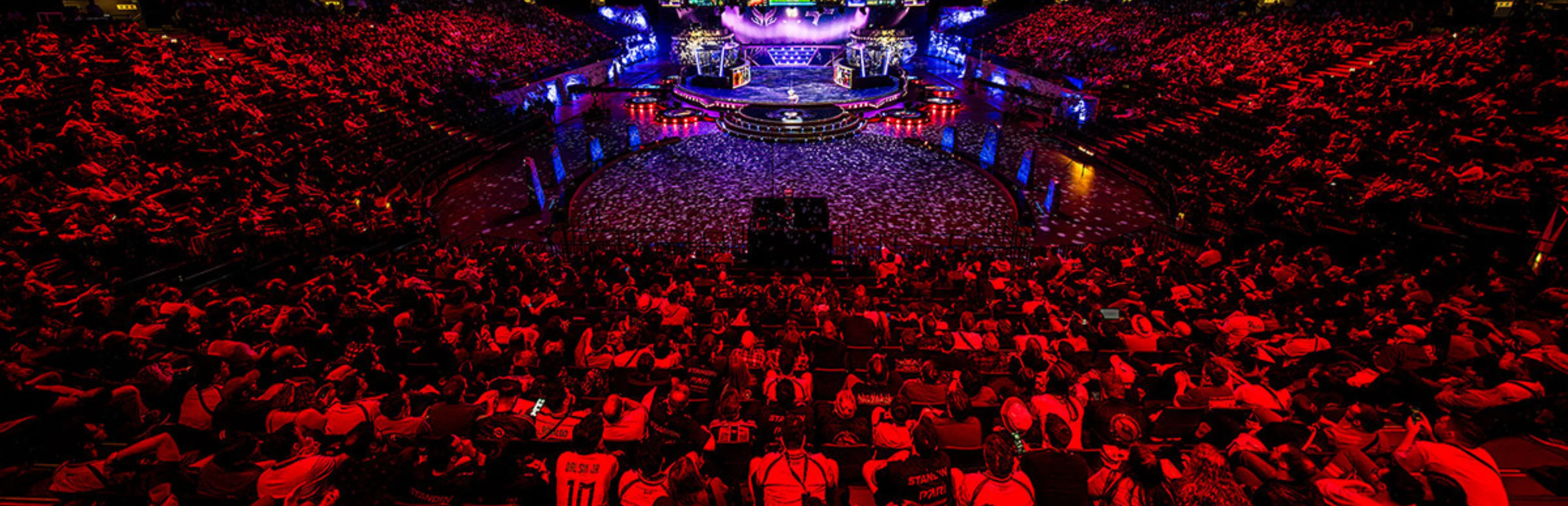 Large esports tournament arena filled with spectators under red lighting, with an illuminated central stage.
