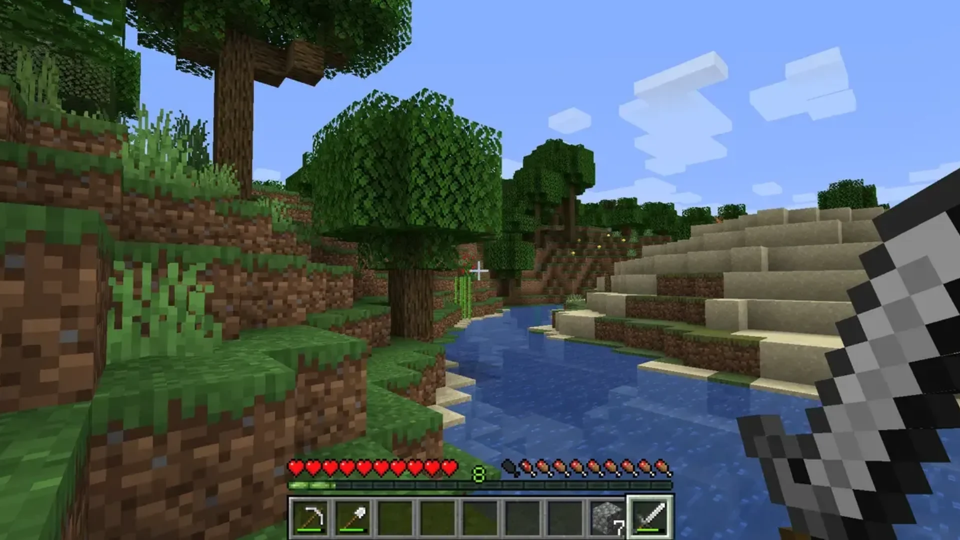 Minecraft Java Edition first-person gameplay beside a river with trees, sand, and an iron sword equipped.